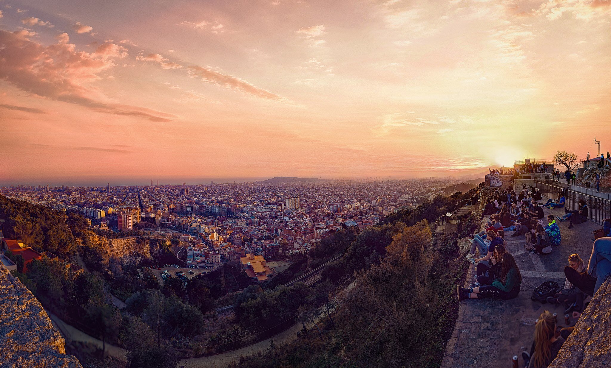 A panoramic view of a city during sunset with a crowd of people sitting on a hilltop overlooking the city skyline and a colorful sky.