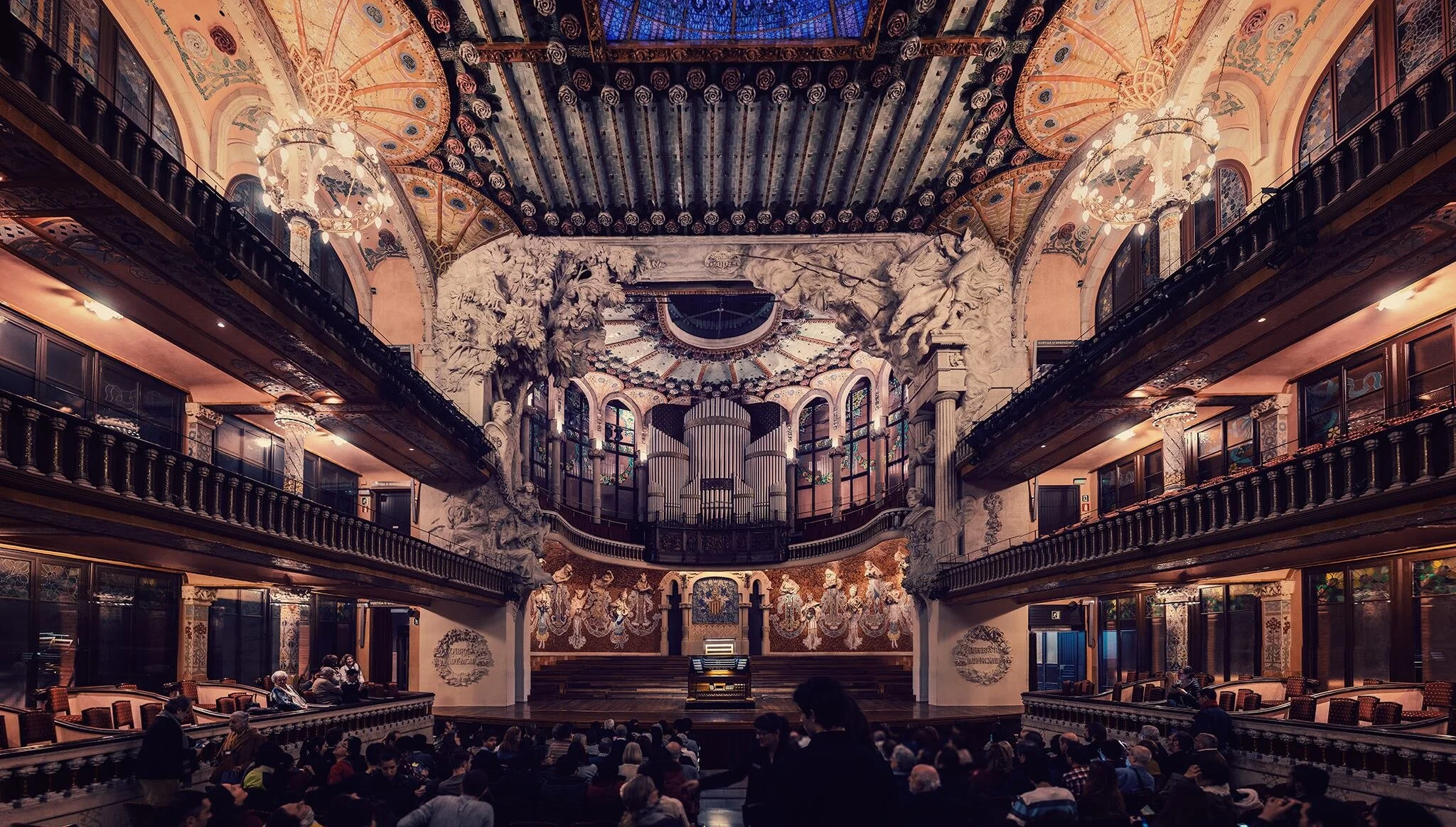 Interior of a grand, ornate concert hall with a stage, elaborate sculptures, and an organ in the center, filled with an audience preparing for a performance.