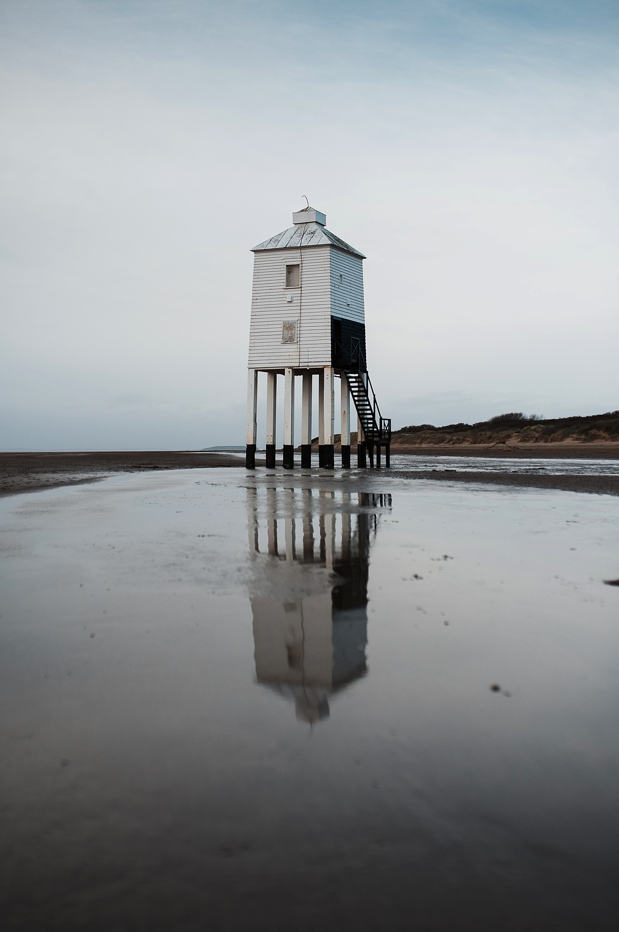 A white wooden lighthouse on stilts standing in shallow water with its reflection visible on the wet surface, under a cloudy sky.
