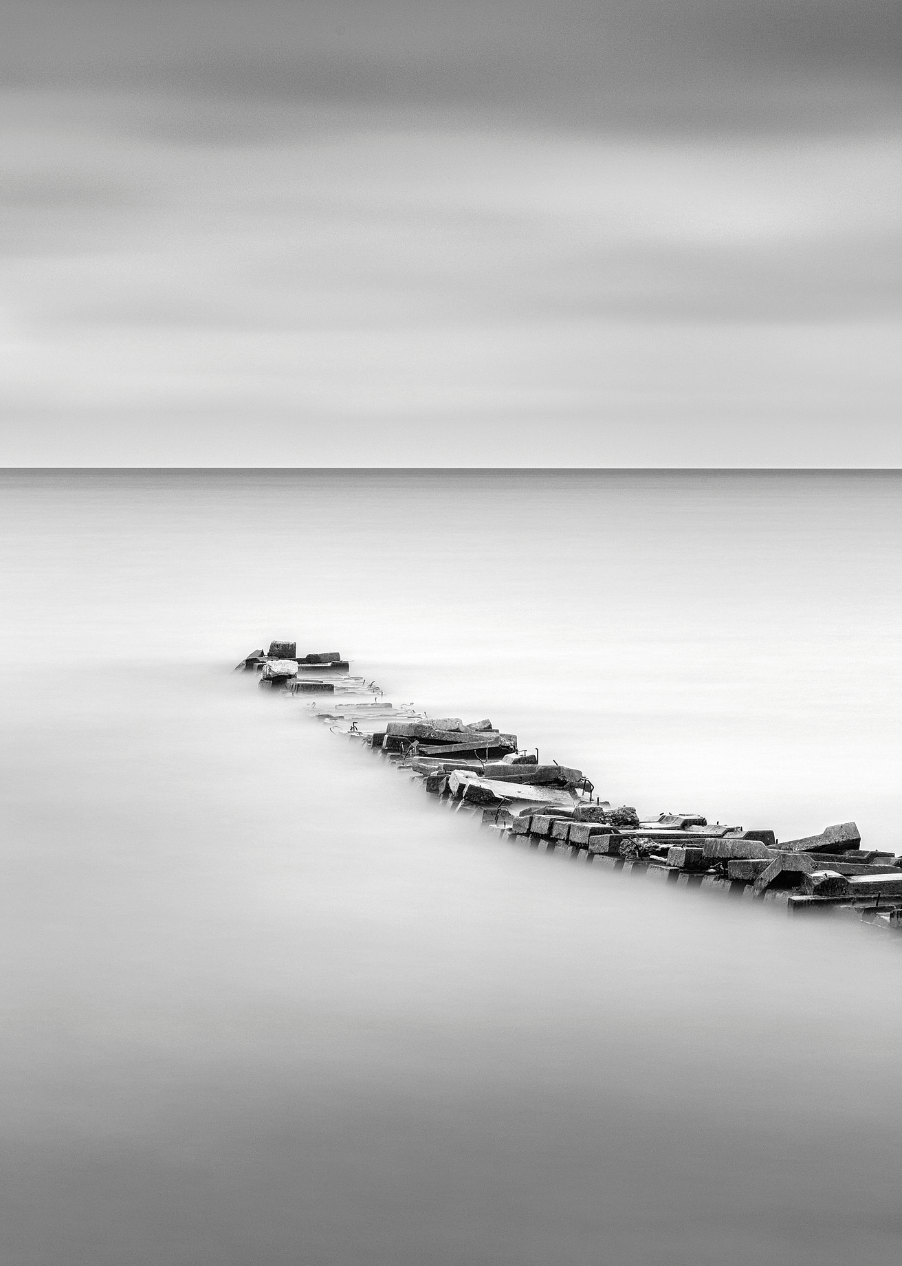 Black and white seascape with a partially submerged broken wooden pier extending into calm water under a cloudy sky.