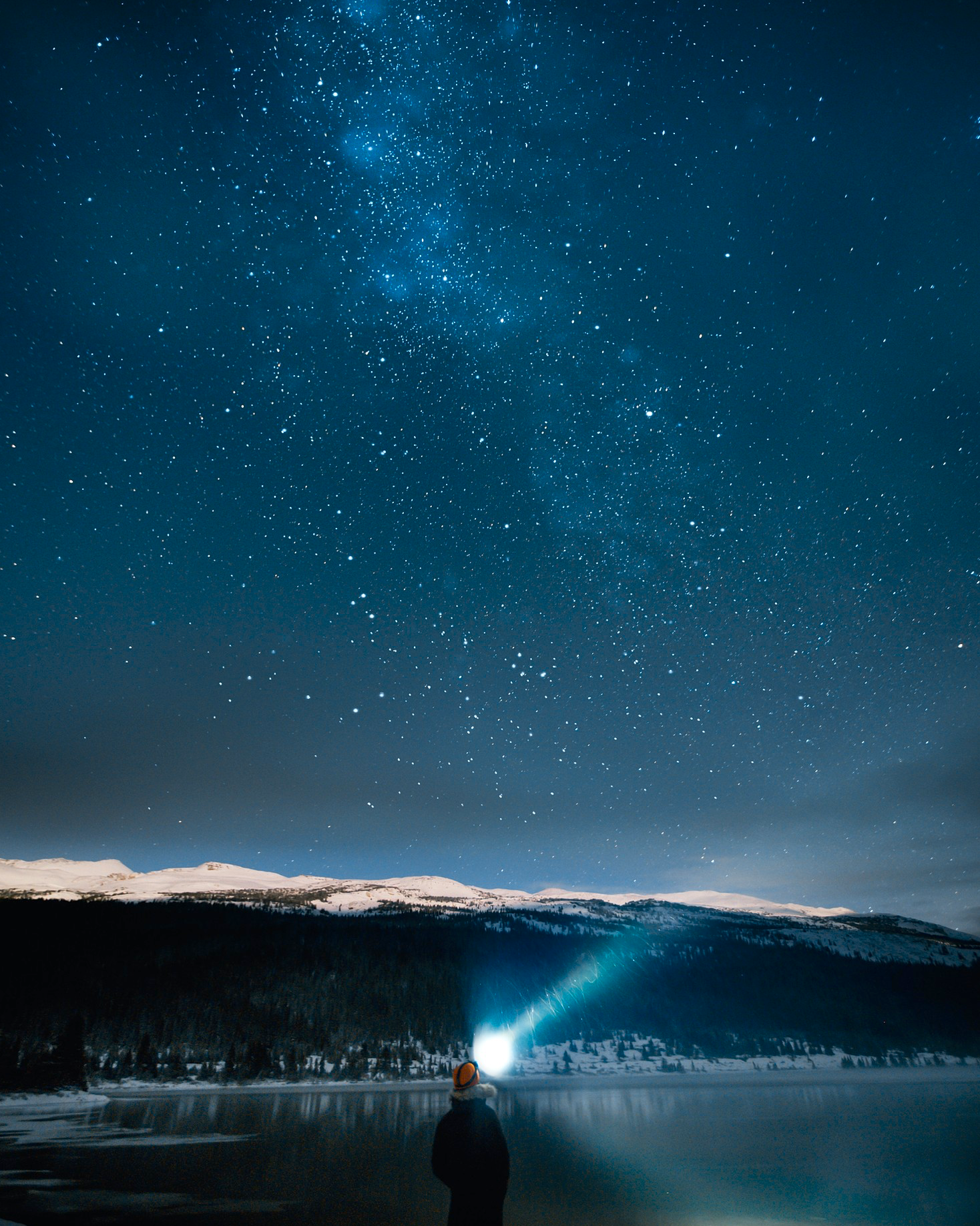A person standing by a lake at night, pointing a flashlight at the star-filled sky and Milky Way galaxy.
