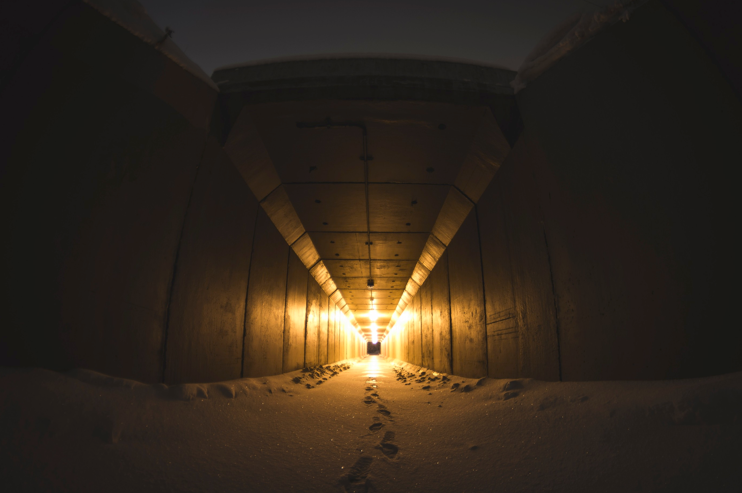 Snow-covered ground inside a tunnel with illuminated lights at the far end, taken from inside looking outward.