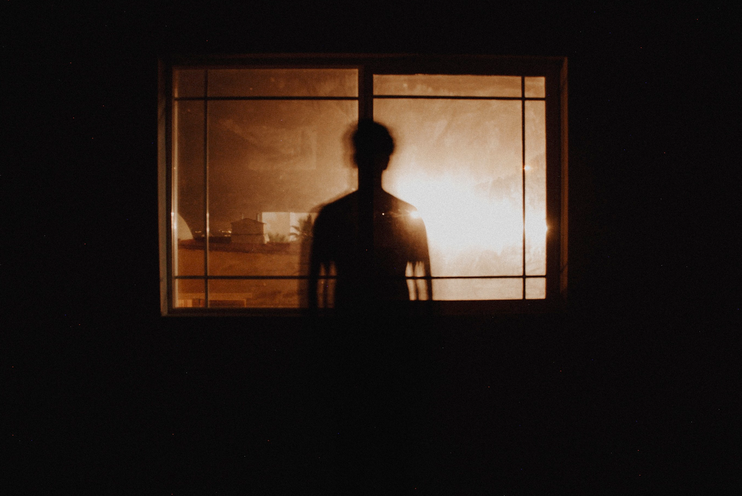 A person standing in front of a lit window at night, with their silhouette visible, inside a dark room.