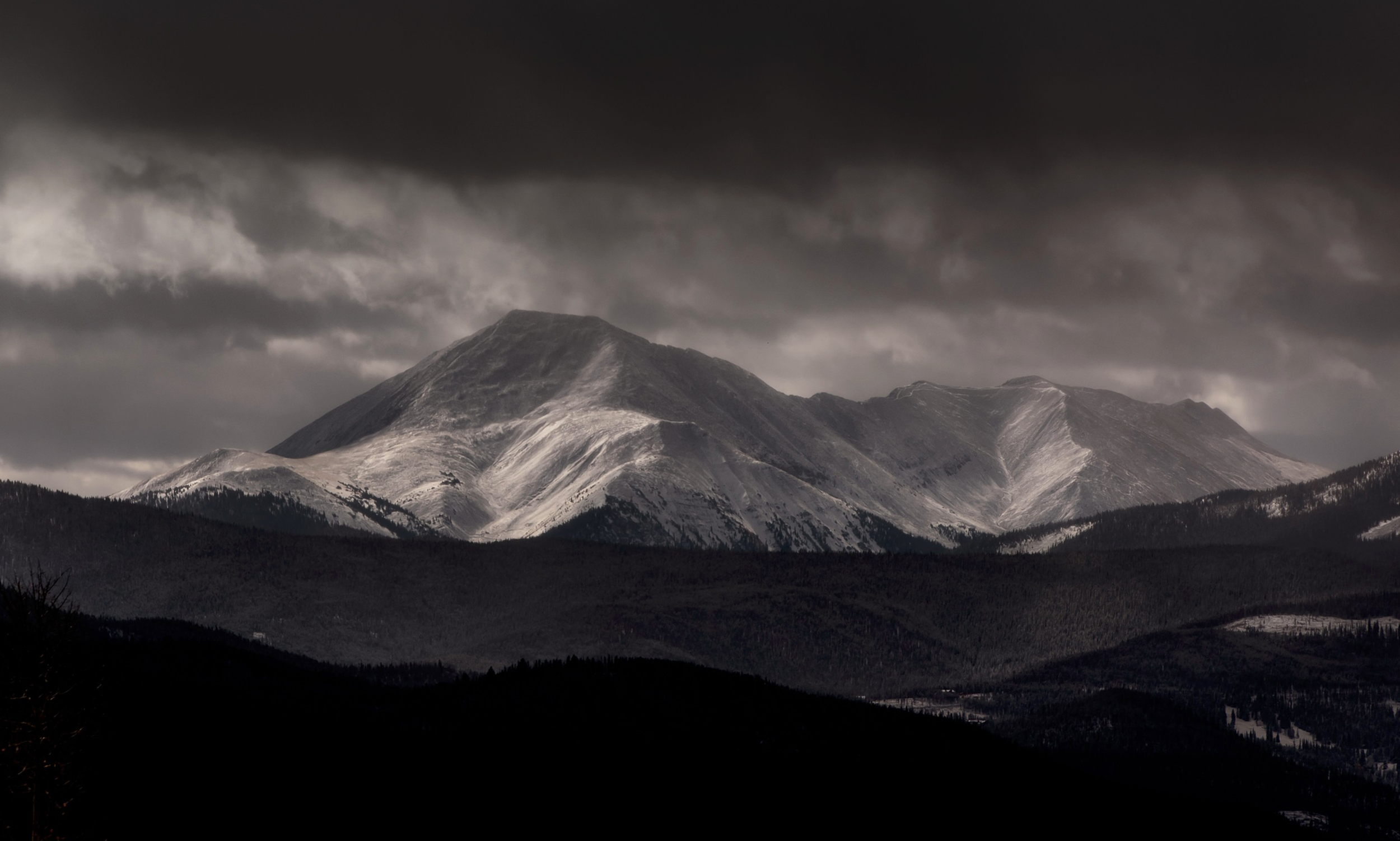Dark mountain range with snow-capped peaks under ominous cloudy sky, black forested hills in foreground.