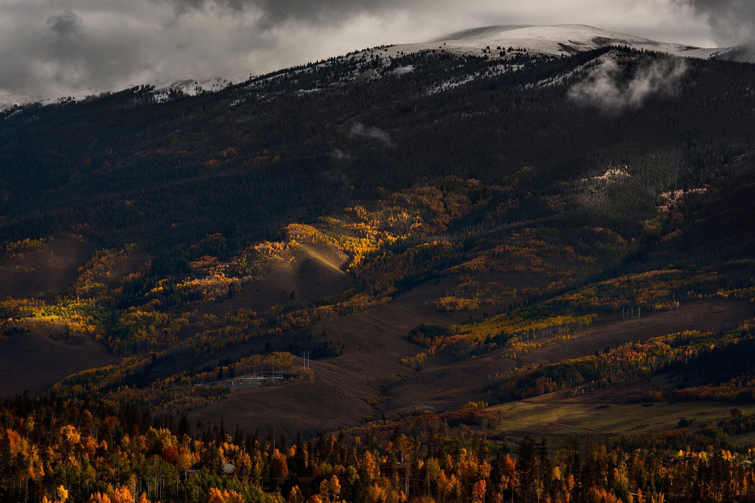 A mountain landscape during autumn with colorful trees, some snow on the mountain peak, and misty clouds.