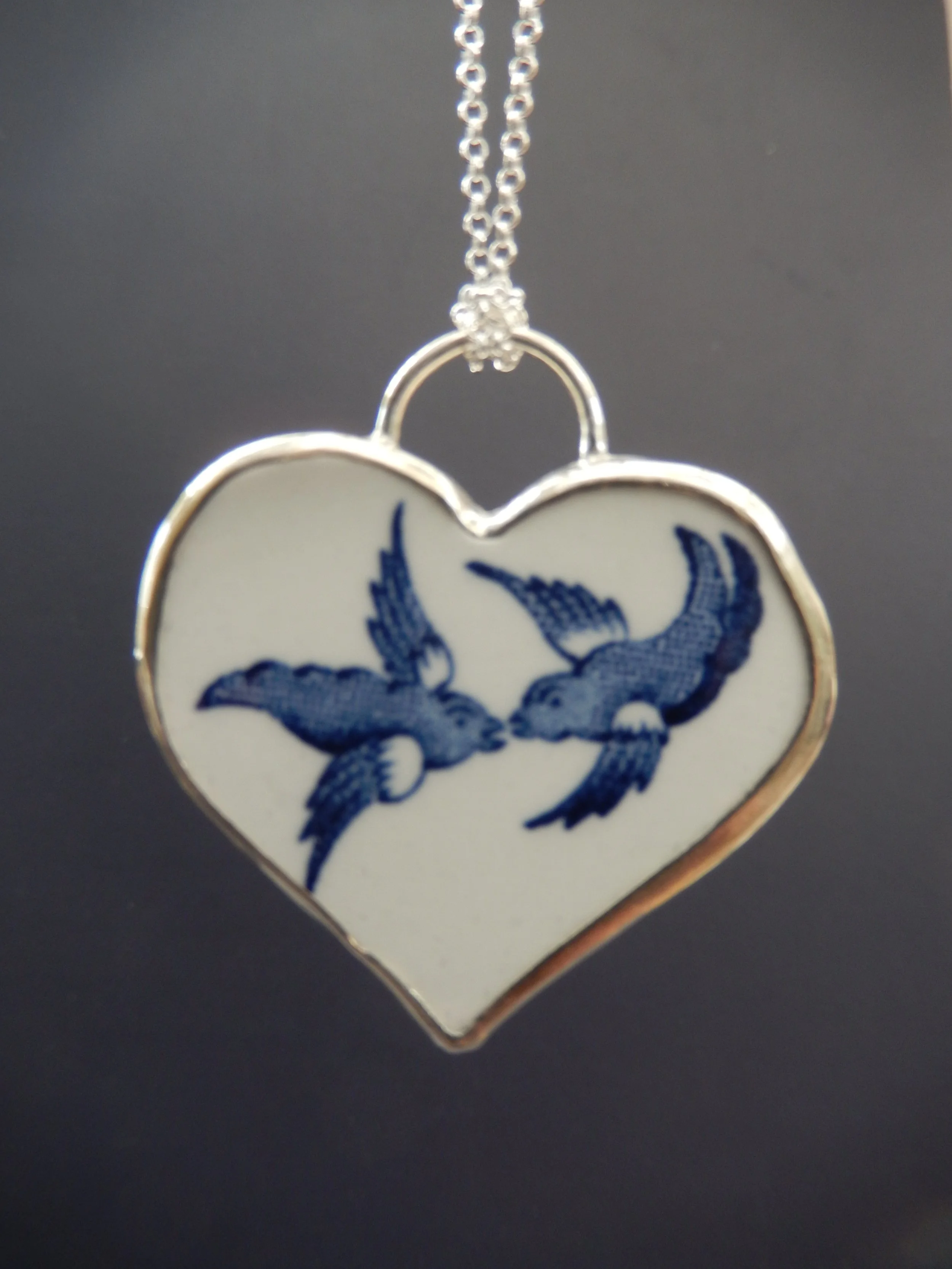 A heart-shaped pendant with blue Willow bird design, hanging from a silver chain. New life to broken plates. Handmade jewellery for 20th wedding anniversary. Handmade in Wales. Unique jewellery.