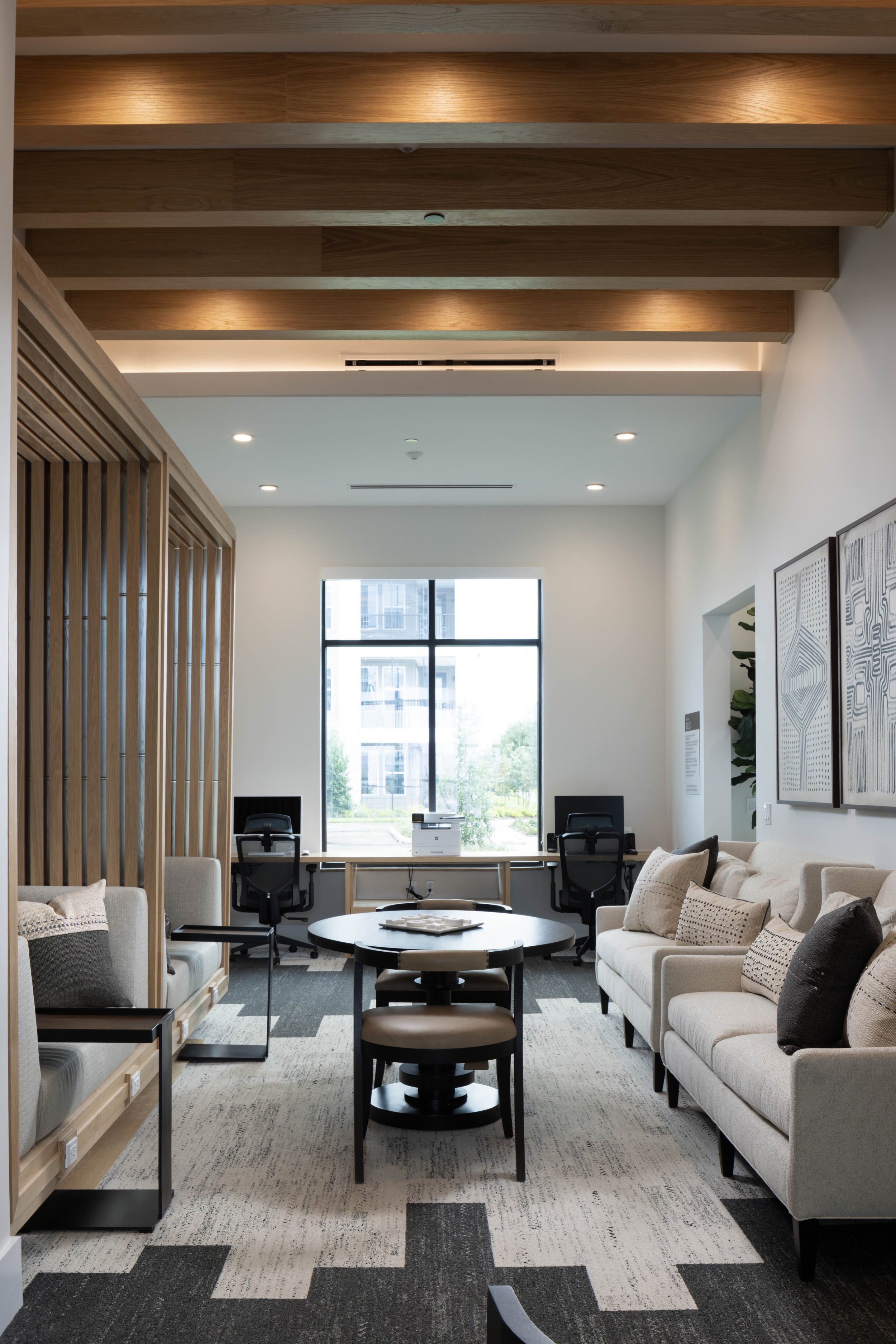 Modern office lounge area with beige sofas, decorative pillows, black chairs, wooden slat room divider, large window with city view, abstract wall art, and patterned carpet.