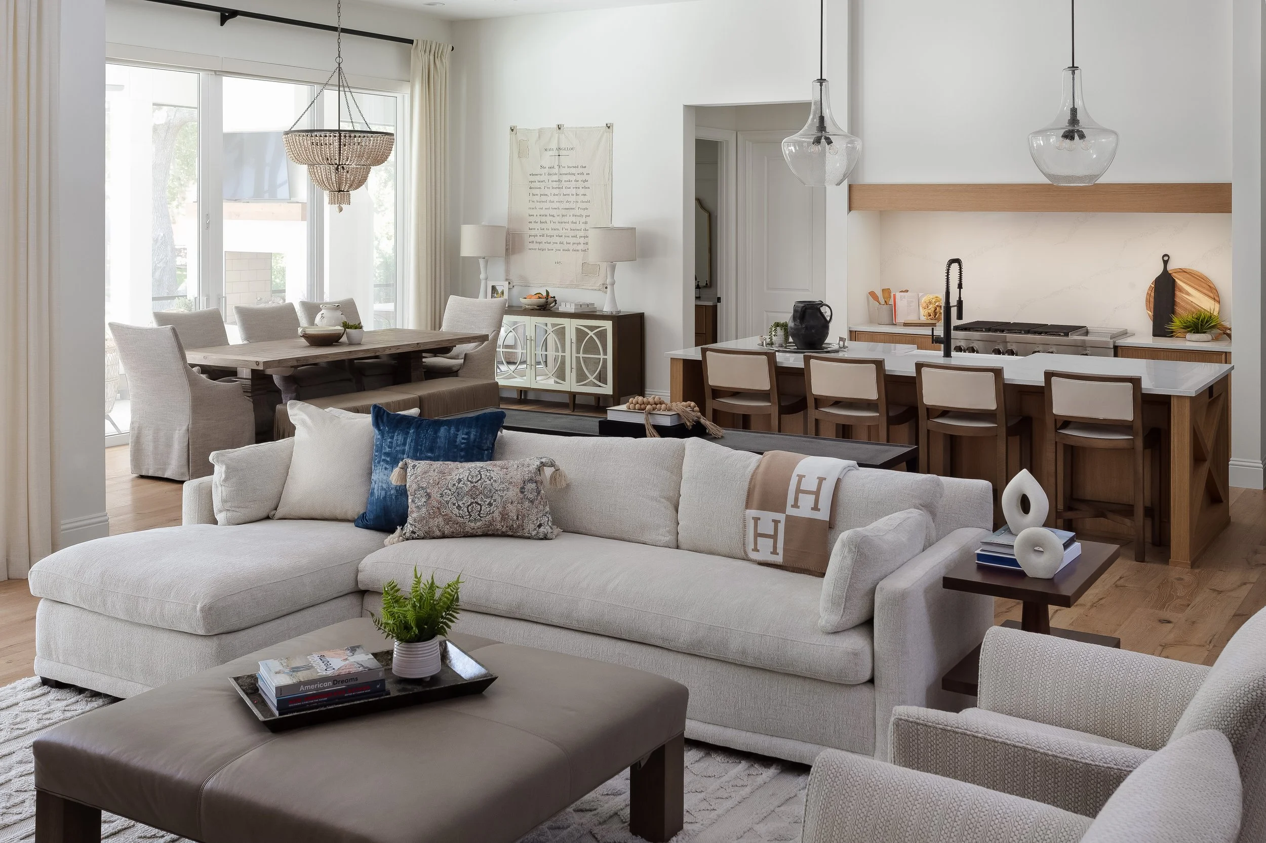 A living room and dining area with neutral tones and modern decor. There is a large white sofa with throw pillows, a wooden dining table with upholstered chairs, and a kitchen with a white counter and natural wood accents. Large windows let in natura