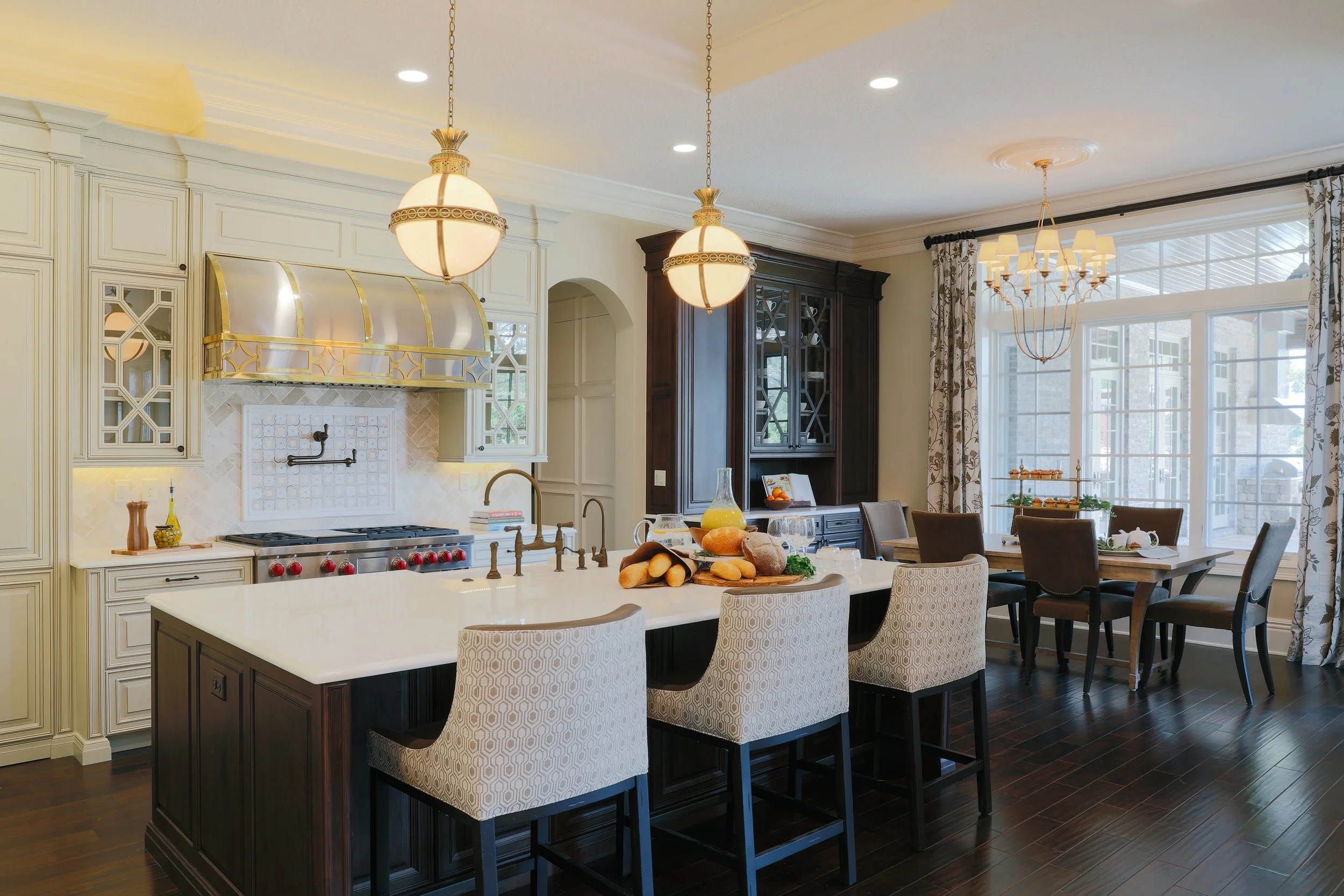 Bright modern kitchen and dining area with large window, white countertops, dark wood cabinetry, hanging lights, and a kitchen island with bread and drinks on top.