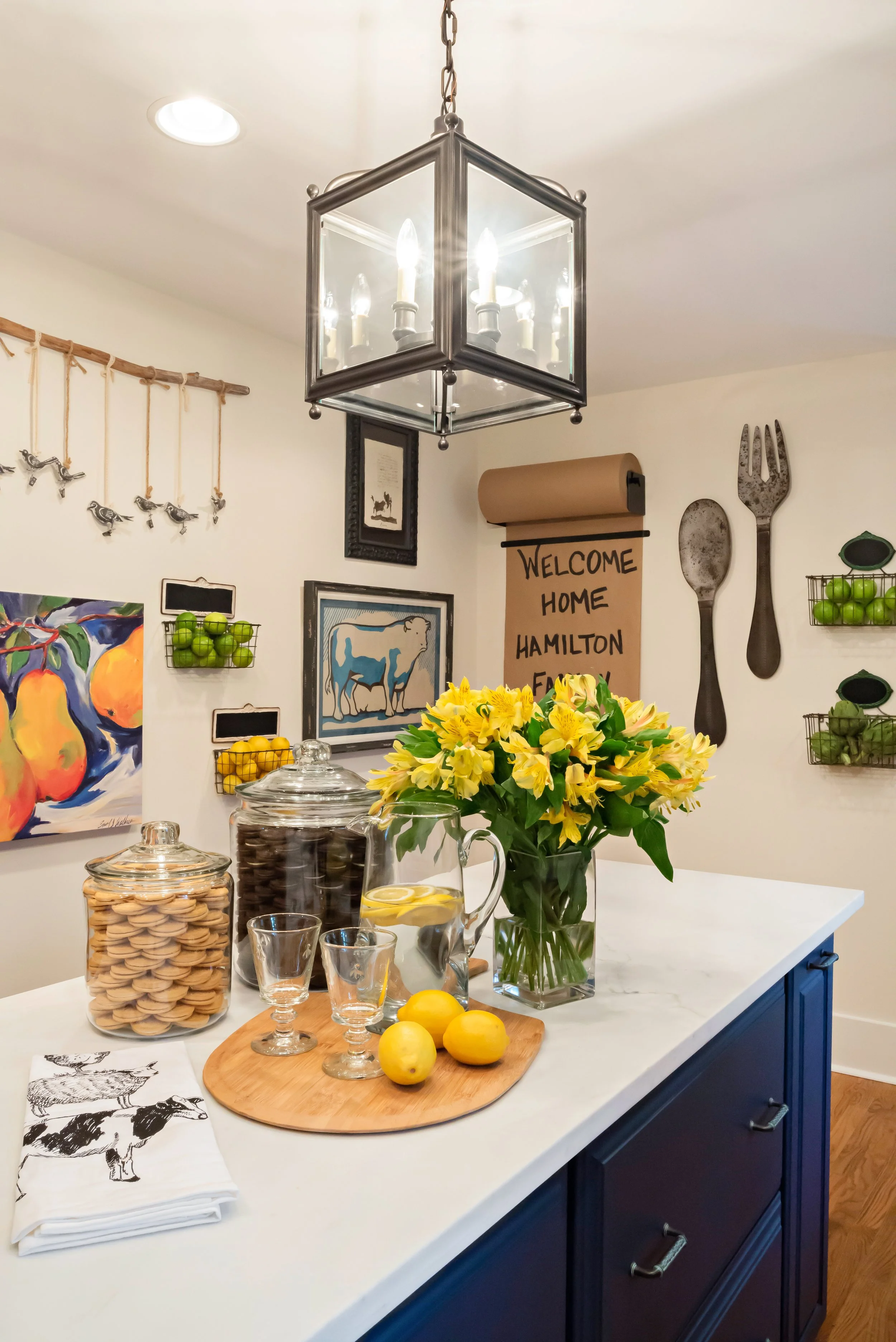 A kitchen island with a floral arrangement of yellow flowers in a glass vase, several glass pitchers with lemon slices, jars of cookies and chocolates, and lemons on a wooden cutting board. The background has colorful artwork, a metal fork and spoon 