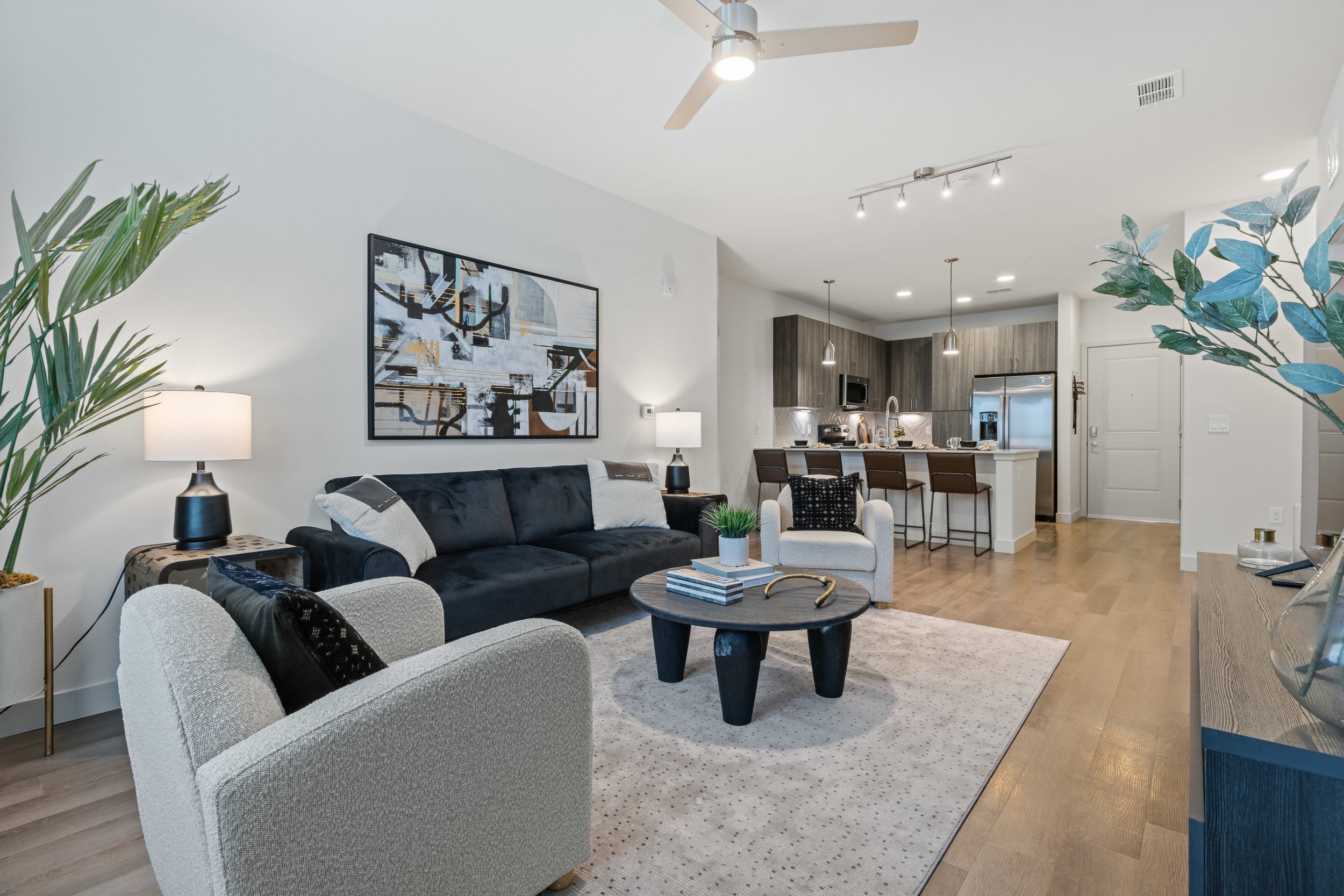 Living room with white walls, black and white couches, and a large abstract art piece on the wall. There are side tables with lamps, a round coffee table with books, and large potted plants. The open kitchen area features a breakfast bar with bar sto