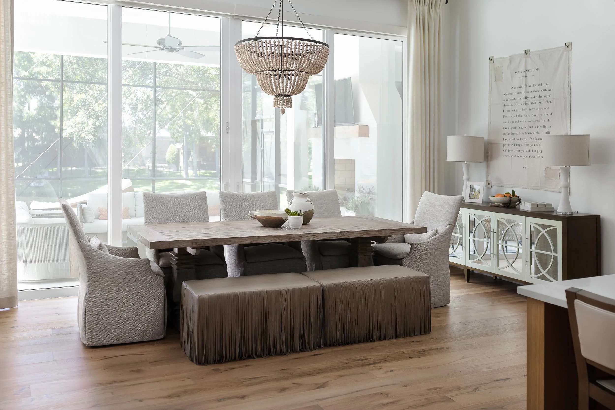 A dining room with a wooden table surrounded by six light gray upholstered chairs. A wrought iron chandelier with beaded decoration hangs above the table. Large sliding glass doors reveal an outdoor patio with a ceiling fan and a view of green trees.