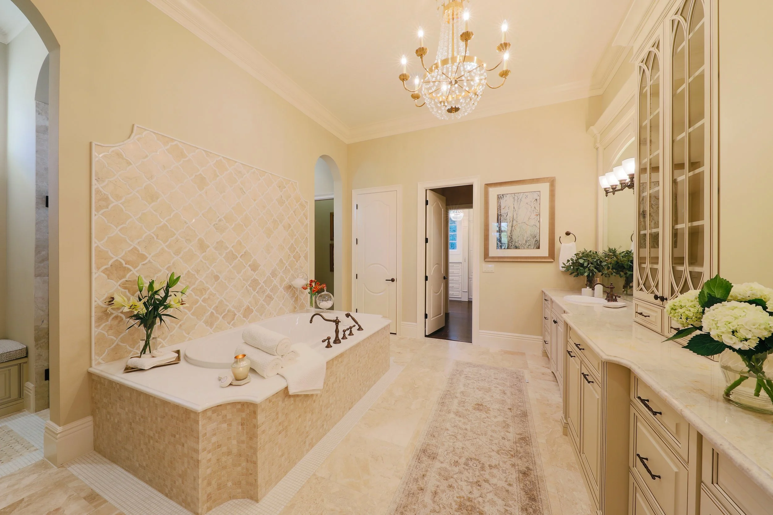 A luxurious bathroom with a chandelier, a bathtub surrounded by beige tiles, and vanity area with flowers and framed artwork.