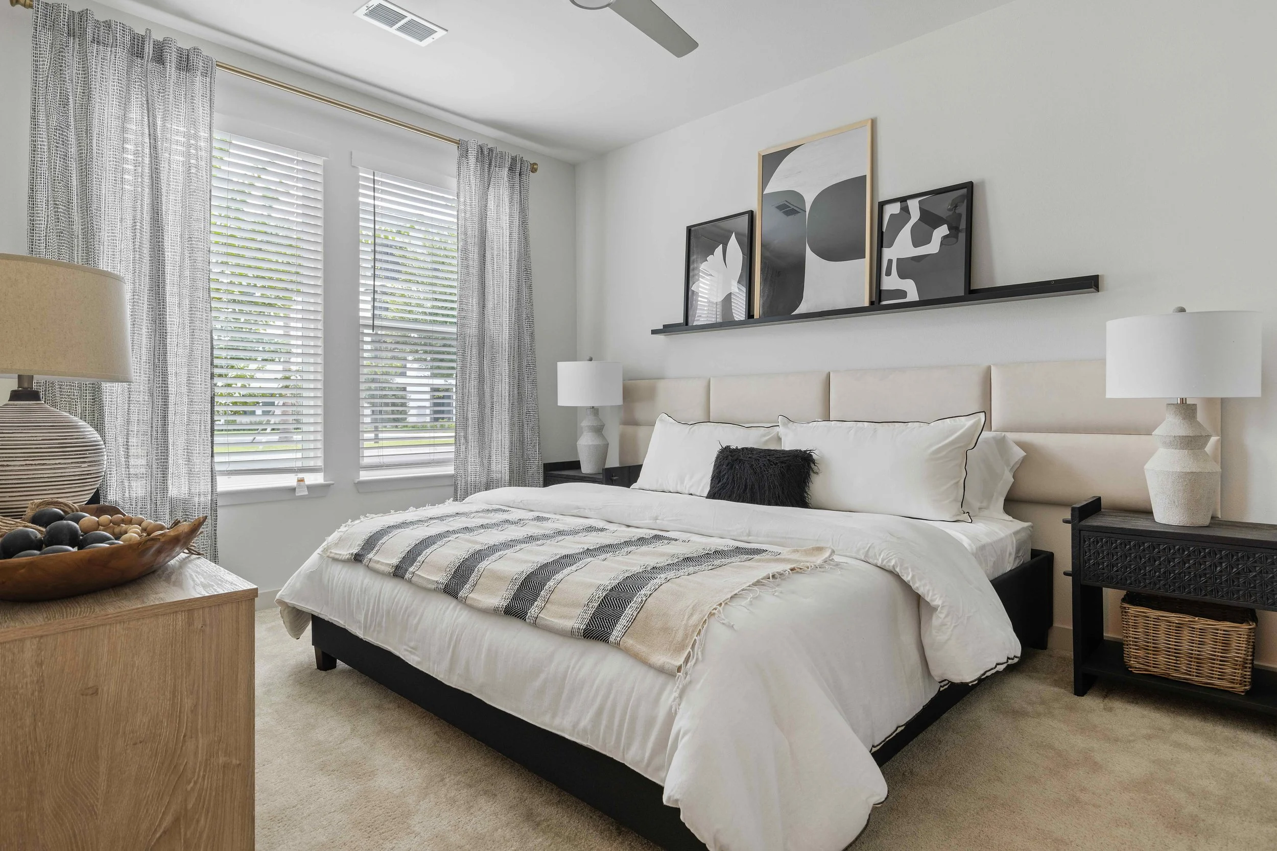 A neatly made bed with white bedding, decorative pillows, and a beige throw blanket in a bright bedroom with large windows, patterned curtains, black nightstands, white lamps, and modern wall art.