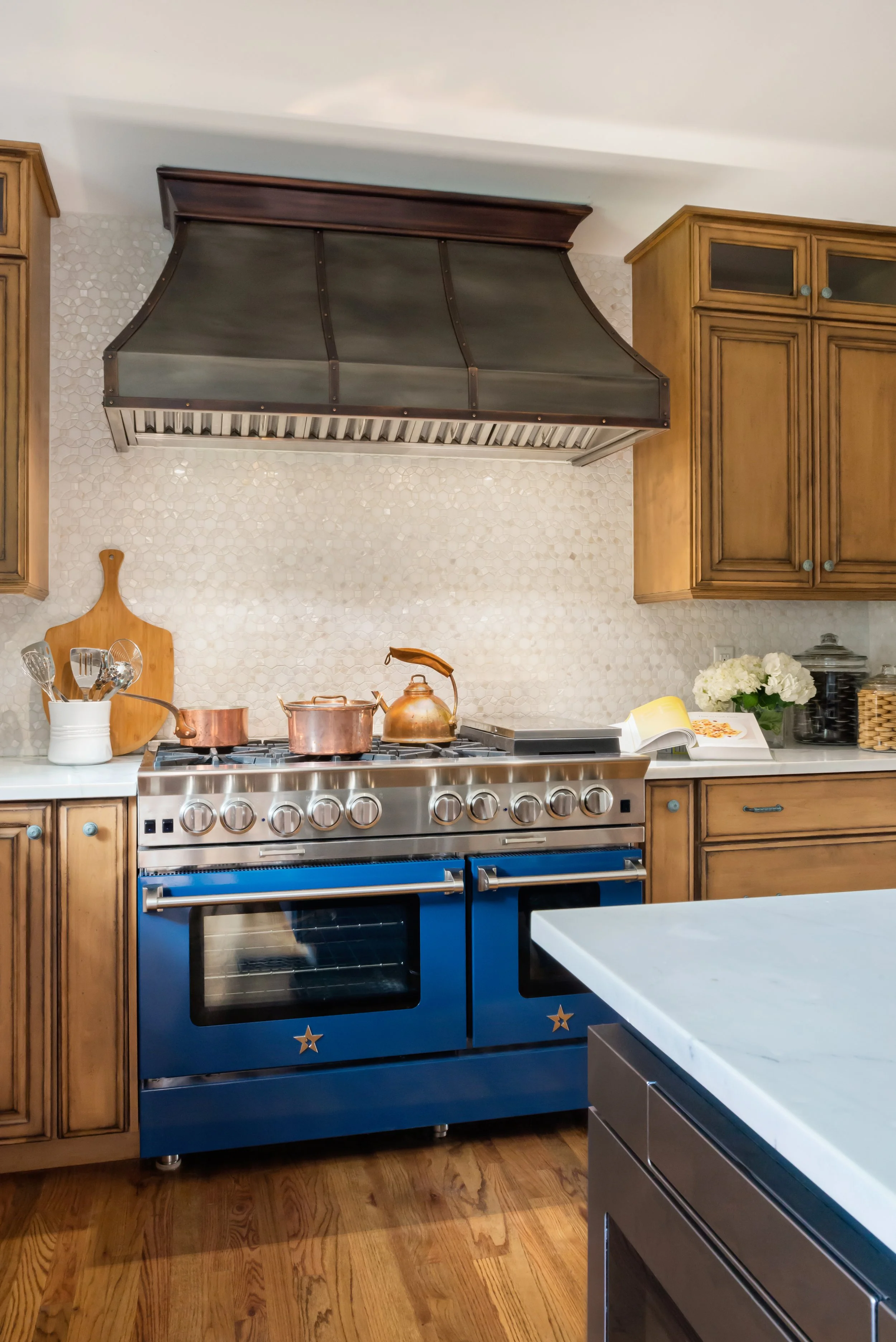 A kitchen with wooden cabinets, a blue stove with star decals, copper pots on the stove, a white marble countertop, and a decorative backsplash with small white tiles. There is a wooden cutting board, white flowers, and kitchen utensils on the counte