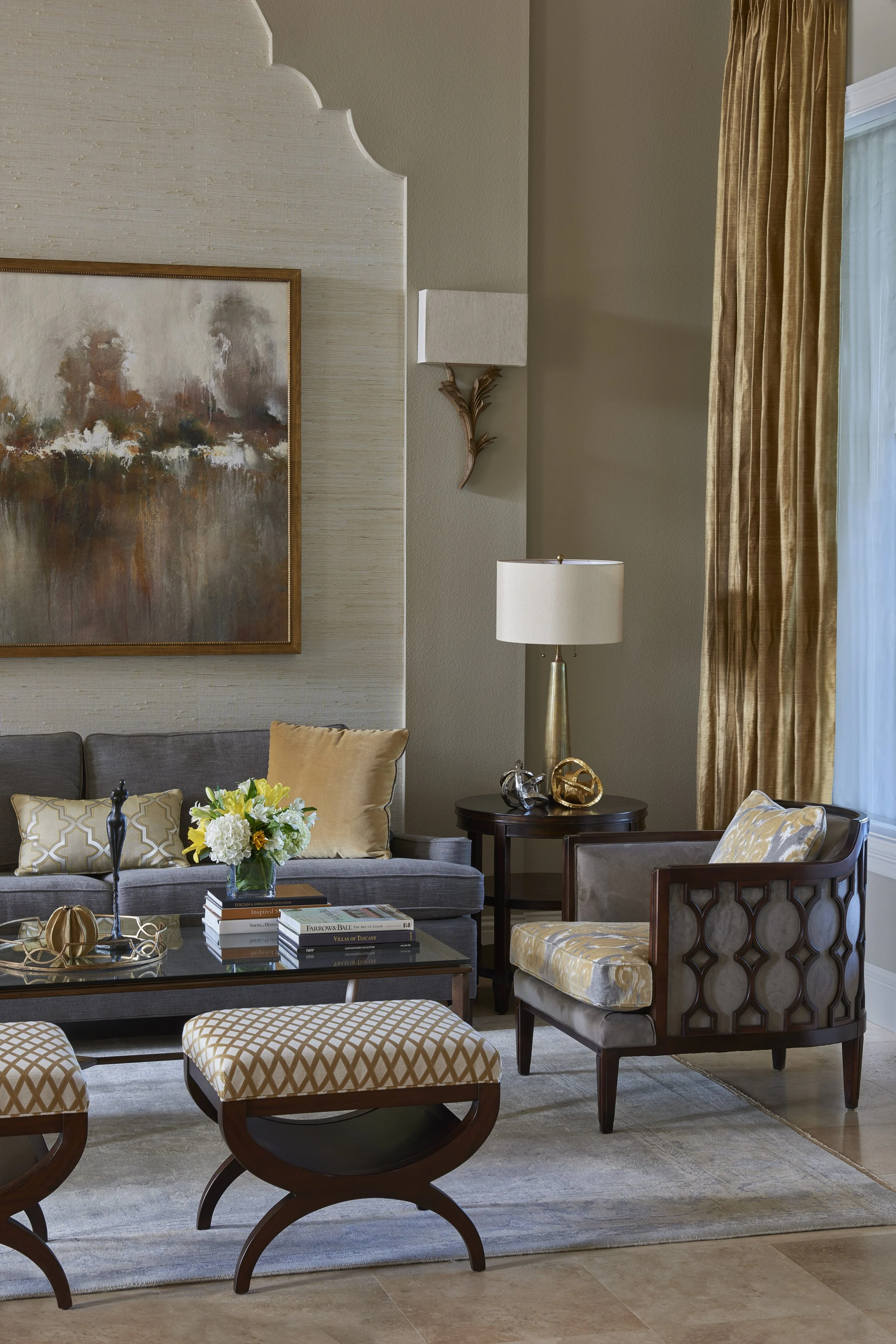 Living room with beige walls, a large third of an abstract painting, a gray sofa with pillows, a glass coffee table with books and a flower arrangement, wood and fabric armchairs, a side table with decorative objects, a gold and white table lamp, and