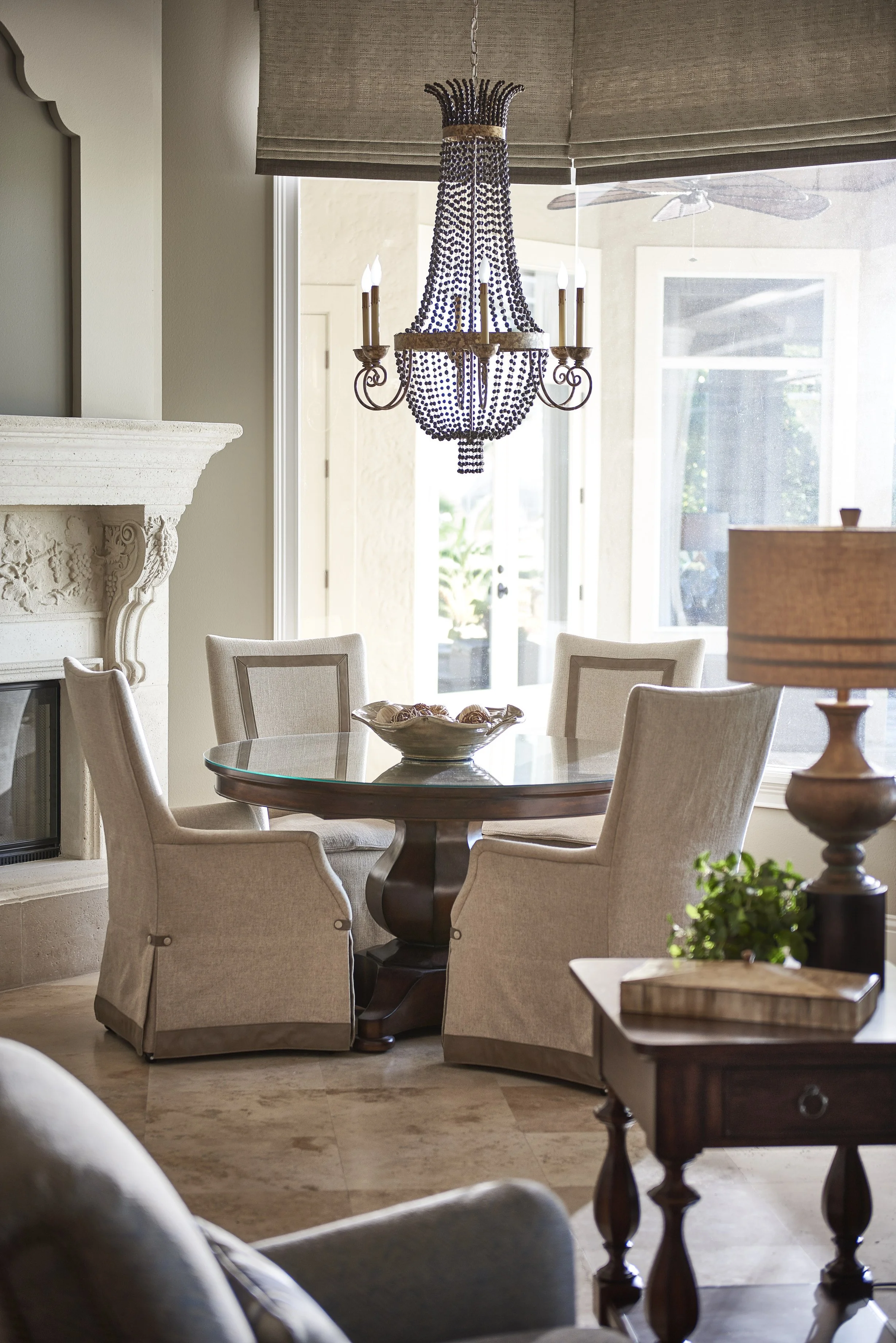 A dining room with a round glass table surrounded by beige upholstered chairs. A decorative bowl with pinecones is on the table. Elegant chandelier with beads hangs above. Large windows let in natural light, with a fan visible outside. Cozy interior 