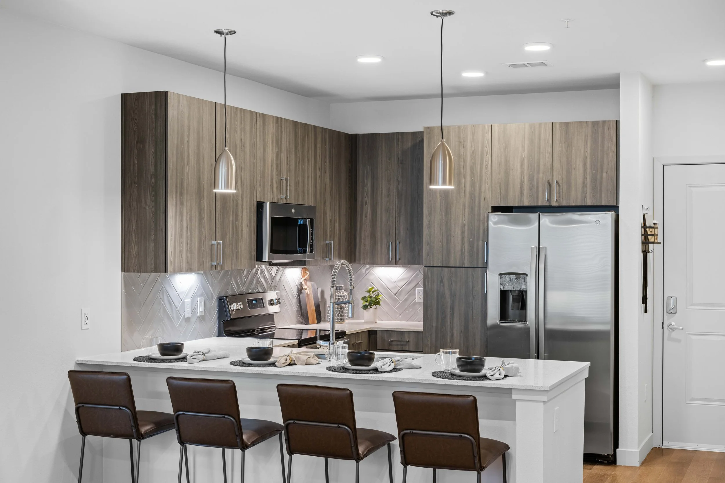 Modern kitchen with wood cabinets, stainless steel appliances, hanging pendant lights, a white island with place settings, and a white wall with a door.