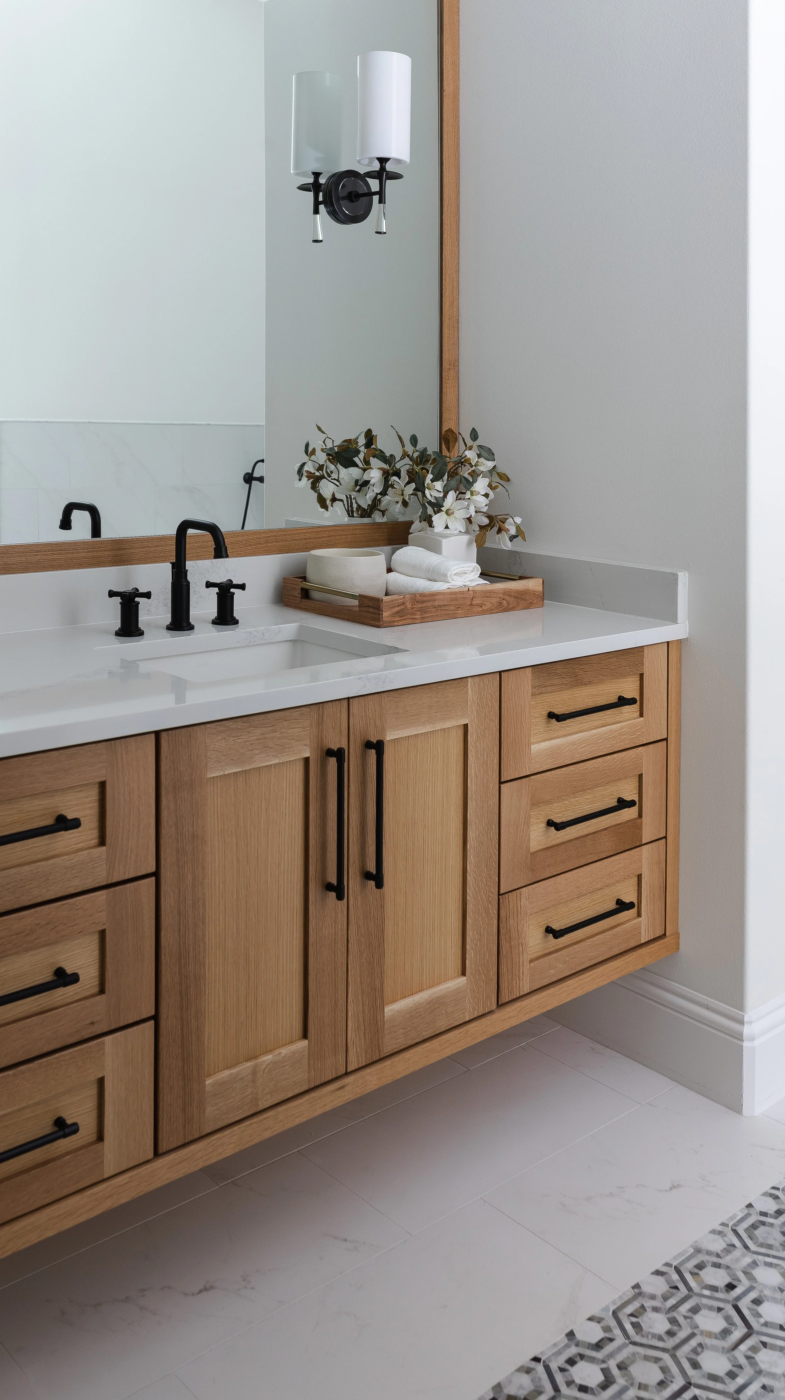Bathroom vanity with wooden cabinets, black hardware, white marble countertop, black faucet, decorative tray with rolled towels and flowers, large mirror, wall sconce light fixture, and tiled floor with patterned border.
