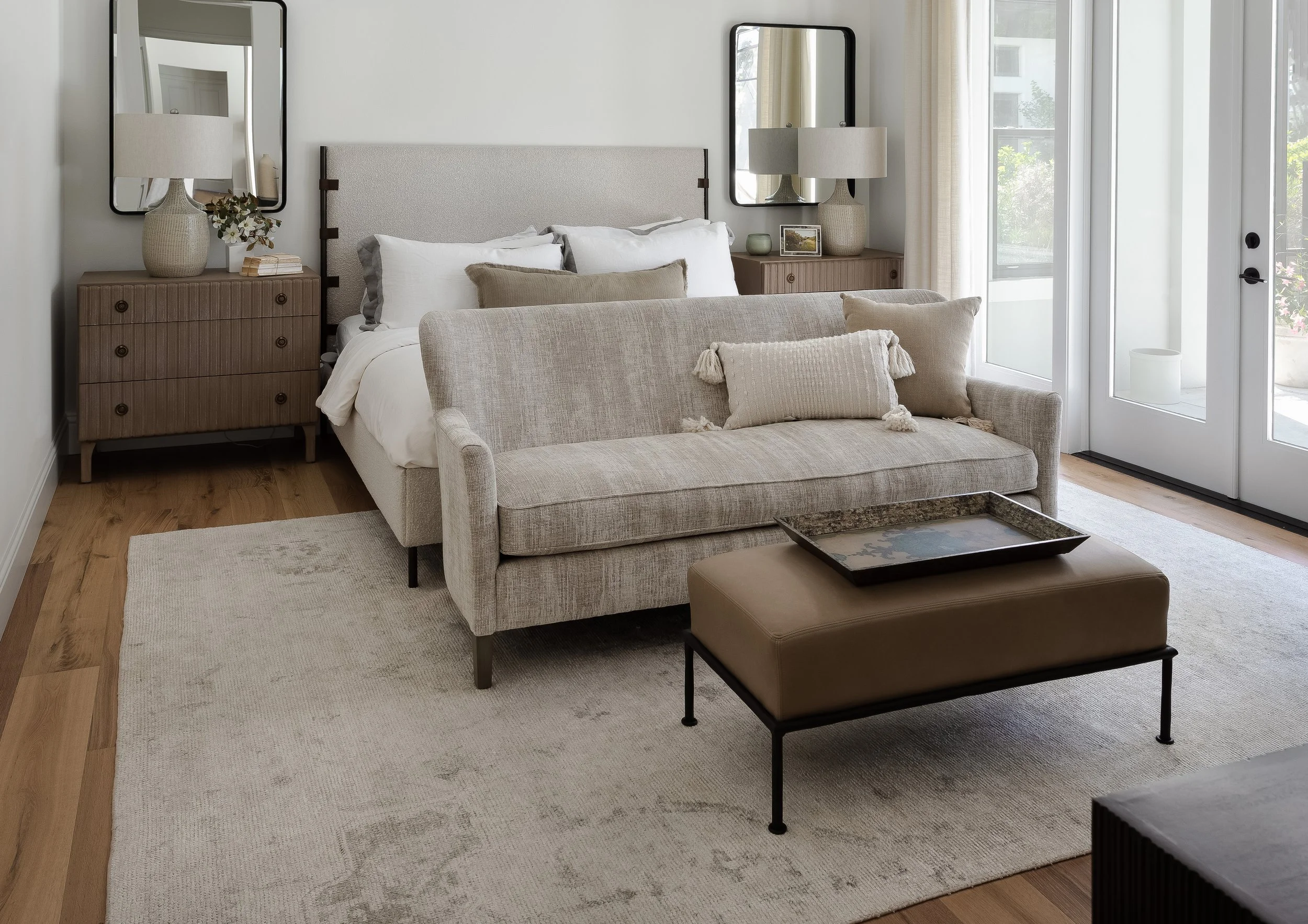 A cozy living room with a beige sofa, decorative pillows, two wooden side tables with lamps, and a large window with curtains letting in natural light.