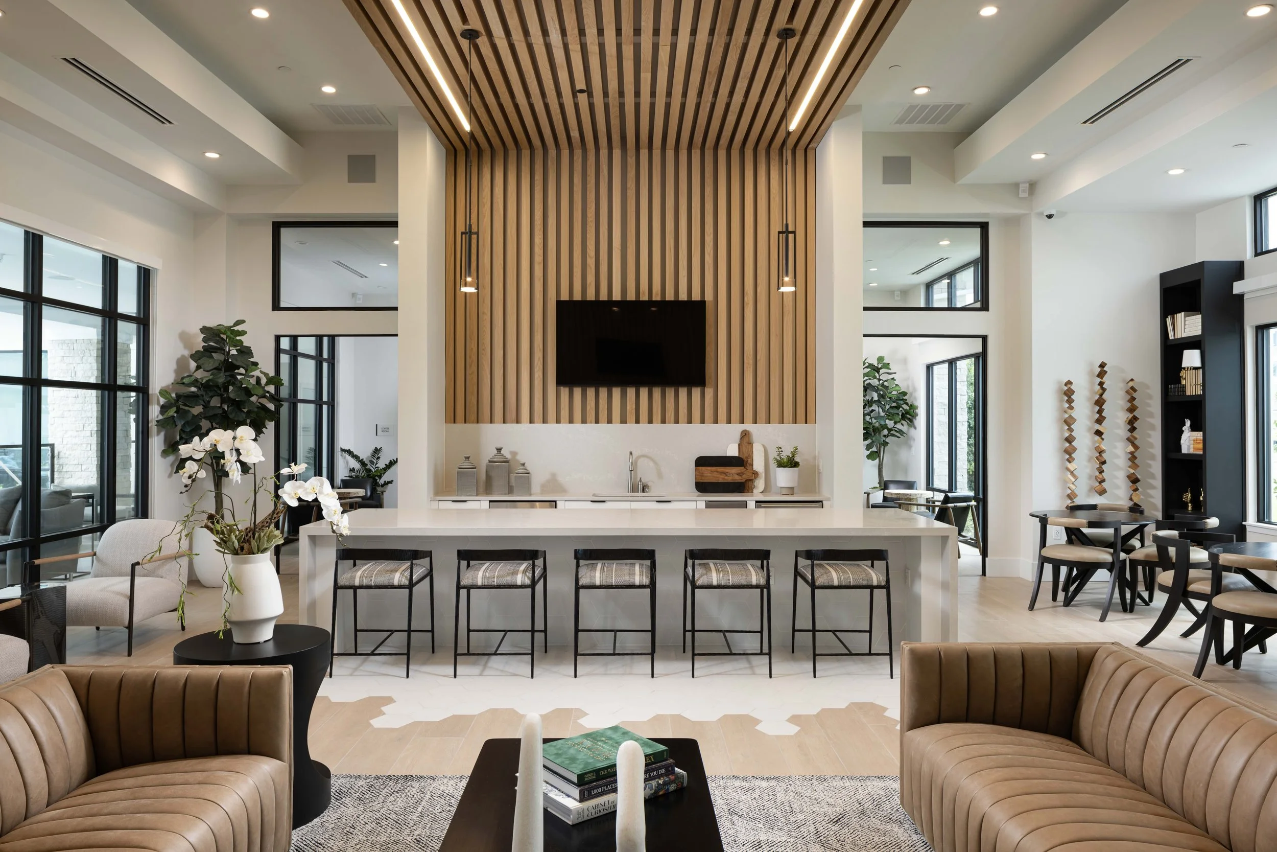 Modern open-concept living room with large windows, a wooden slat feature wall with a mounted TV, and various seating areas including leather armchairs and a beige sofa. Decor includes plants, books, and decorative items.