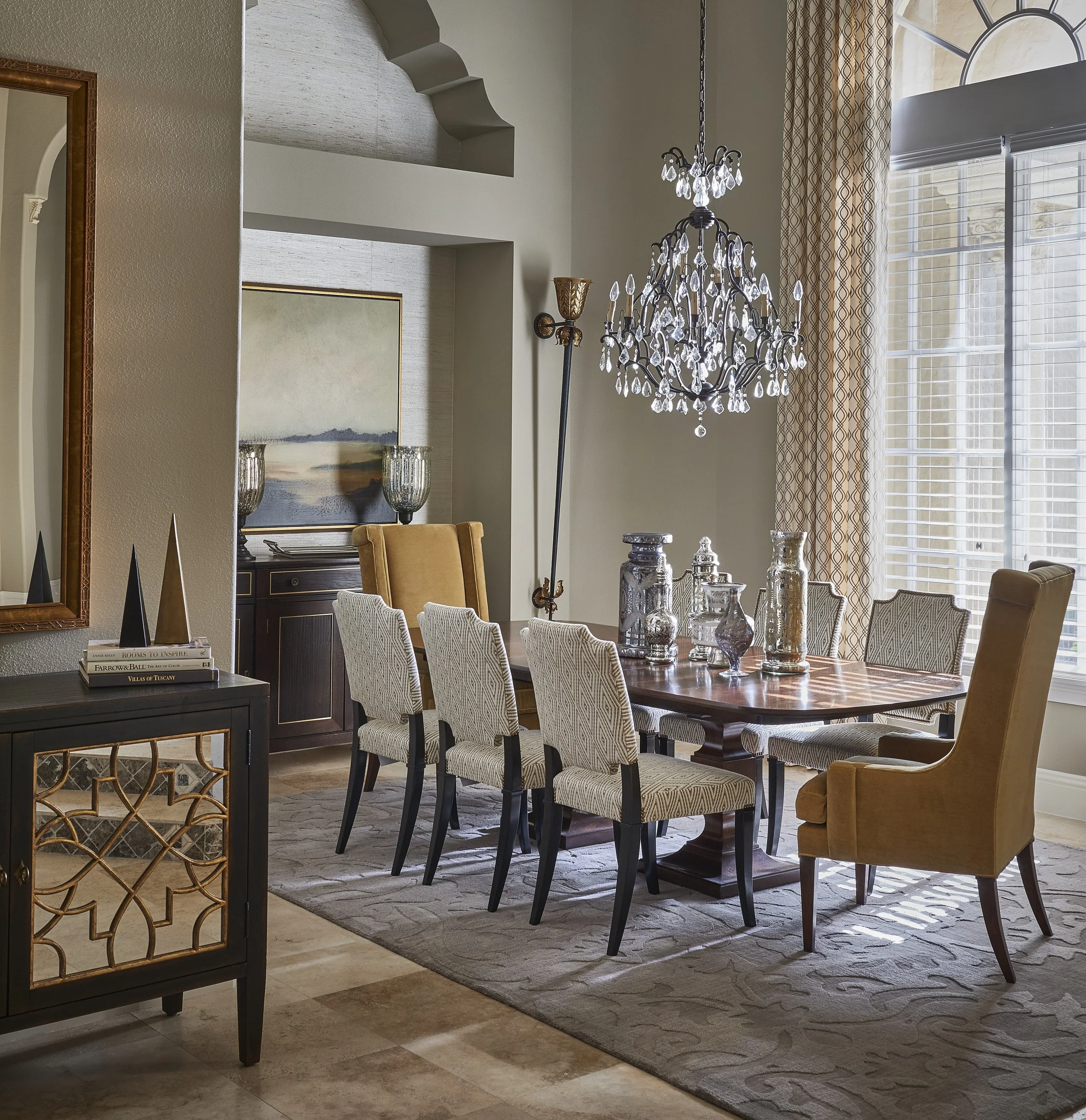 View of a dining room with a large window, a chandelier hanging from the ceiling, a dark wood dining table with beige upholstered chairs, and decorative vases on the table. There is a framed painting on the wall, a sideboard, and patterned curtains.
