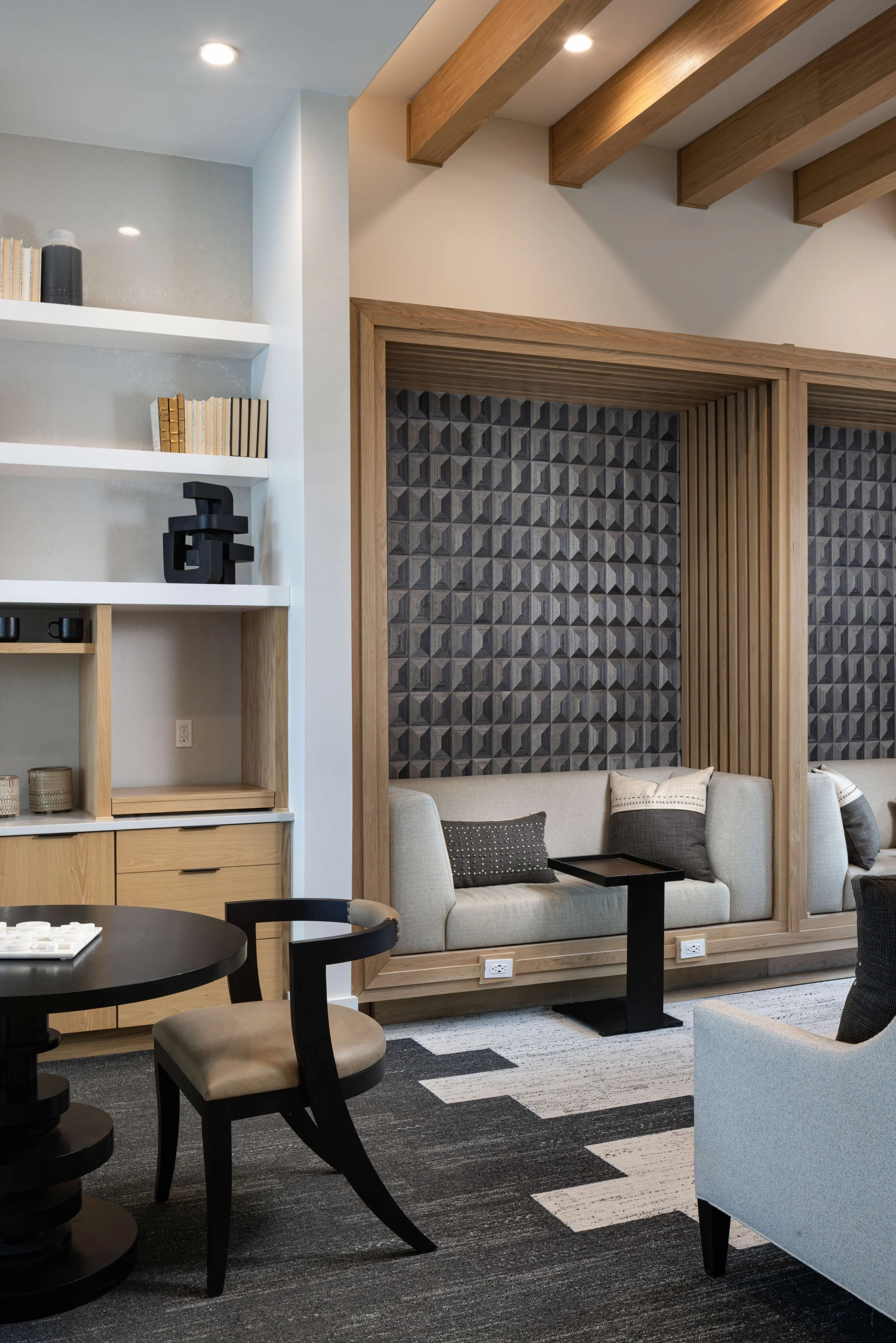 Interior of a modern living room with a beige sofa and decorative pillows, a black side table, a patterned area rug, and wooden accents including ceiling beams and a framed wall with textured tiles. Part of a dining area with a round black table and 