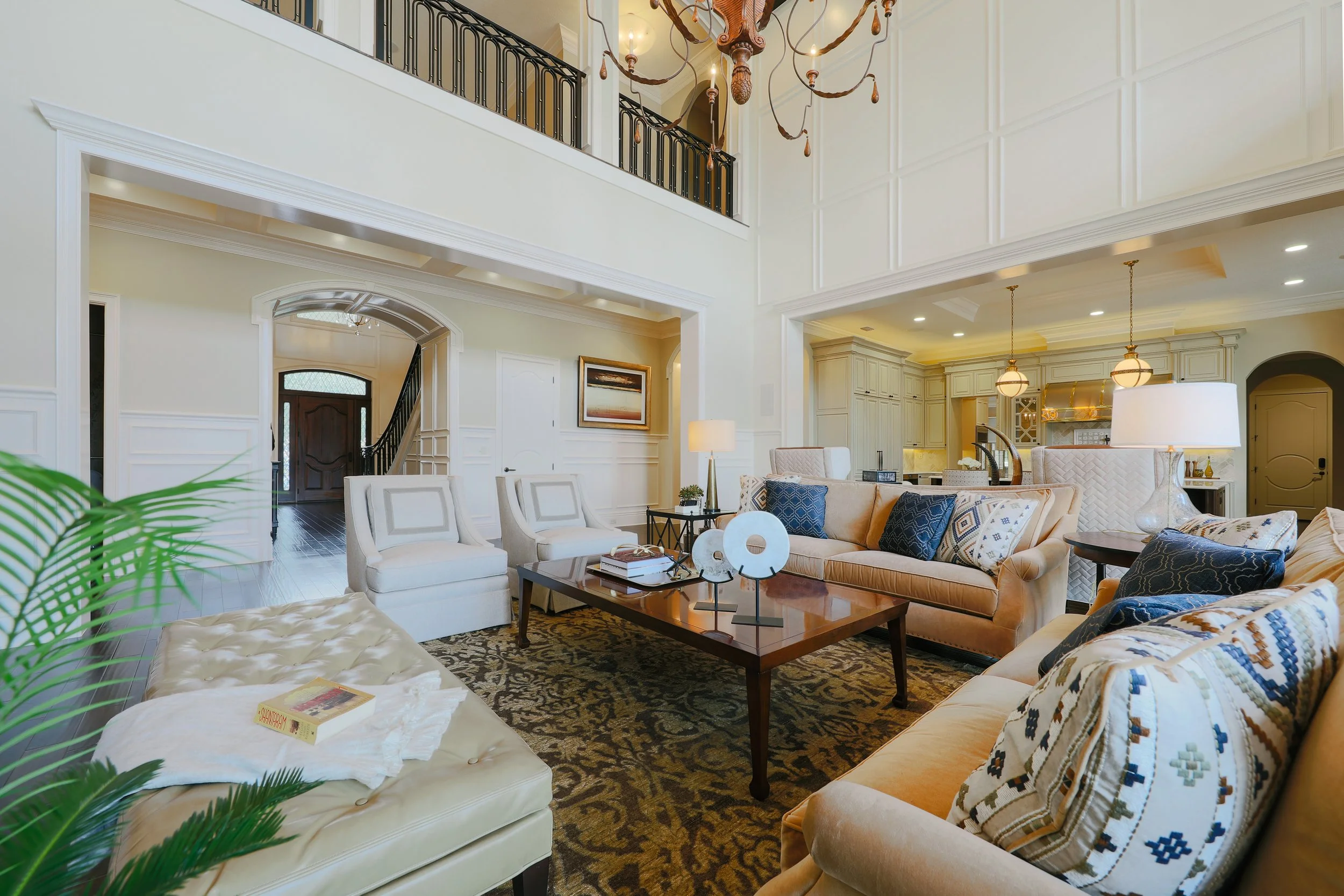 Living room with high ceilings, a chandelier, white walls, and a dark wood staircase. Furniture includes a beige sofa, white armchairs, and a wooden coffee table with decorative items. There are also two hanging pendant lights over a kitchen area wit