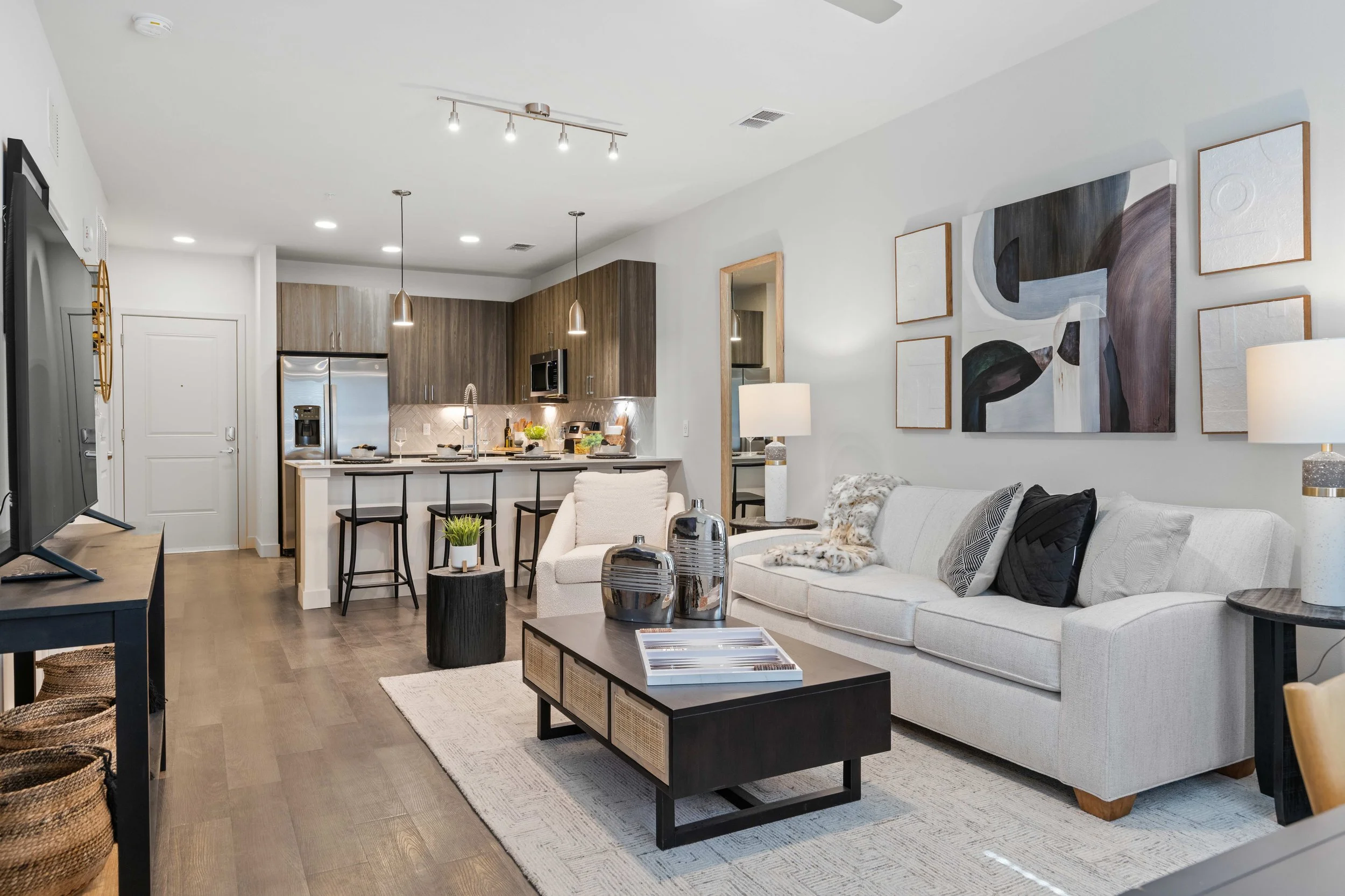 Modern open-plan living room and kitchen with white walls, hardwood floors, a light-colored sofa with black and white pillows, a wooden coffee table, an abstract wall art, and pendant and track lighting.