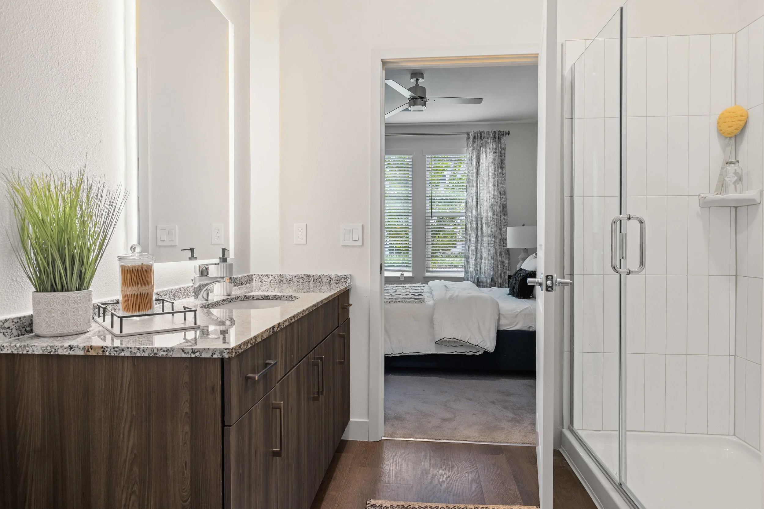 View of a modern bathroom with a granite countertop, a potted plant, and toiletries on the left; a shower enclosure with a handle on the right; and a bedroom with a bed and large windows with blinds visible through an open door in the background.
