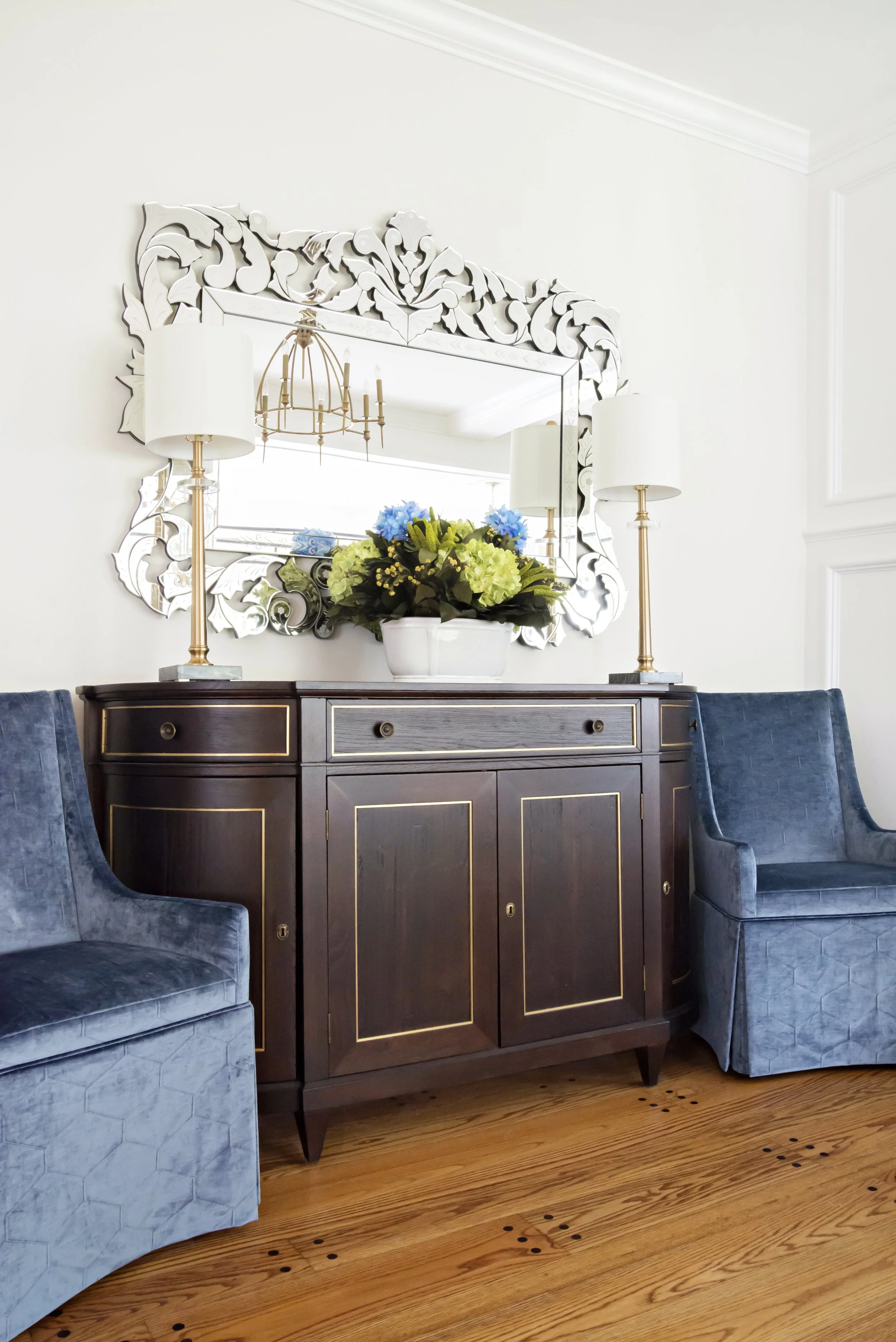 A dark wood sideboard with gold trim and knobs, flanked by two blue velvet armchairs. A white vase with yellow and blue flowers is placed on top of the sideboard. Above, a mirror with an ornate silver frame, a white lampshade, and a gold chandelier a