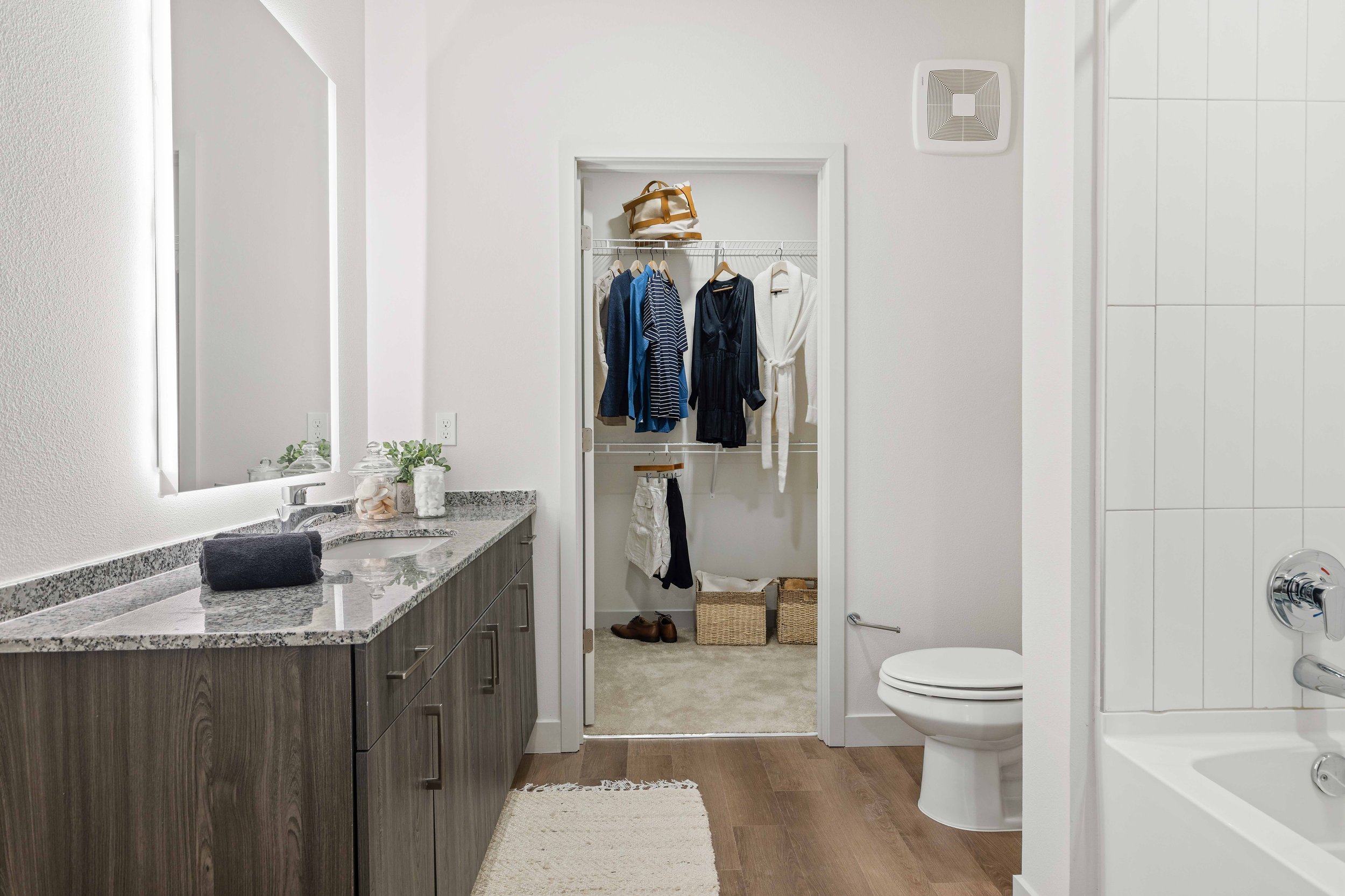 A bathroom with a granite countertop vanity, a mirror, a towel, and a glass container with decorations. An open closet with hanging clothes, shoes, and baskets is visible through a doorway. The bathroom has white walls, a ventilation fan, a toilet, a