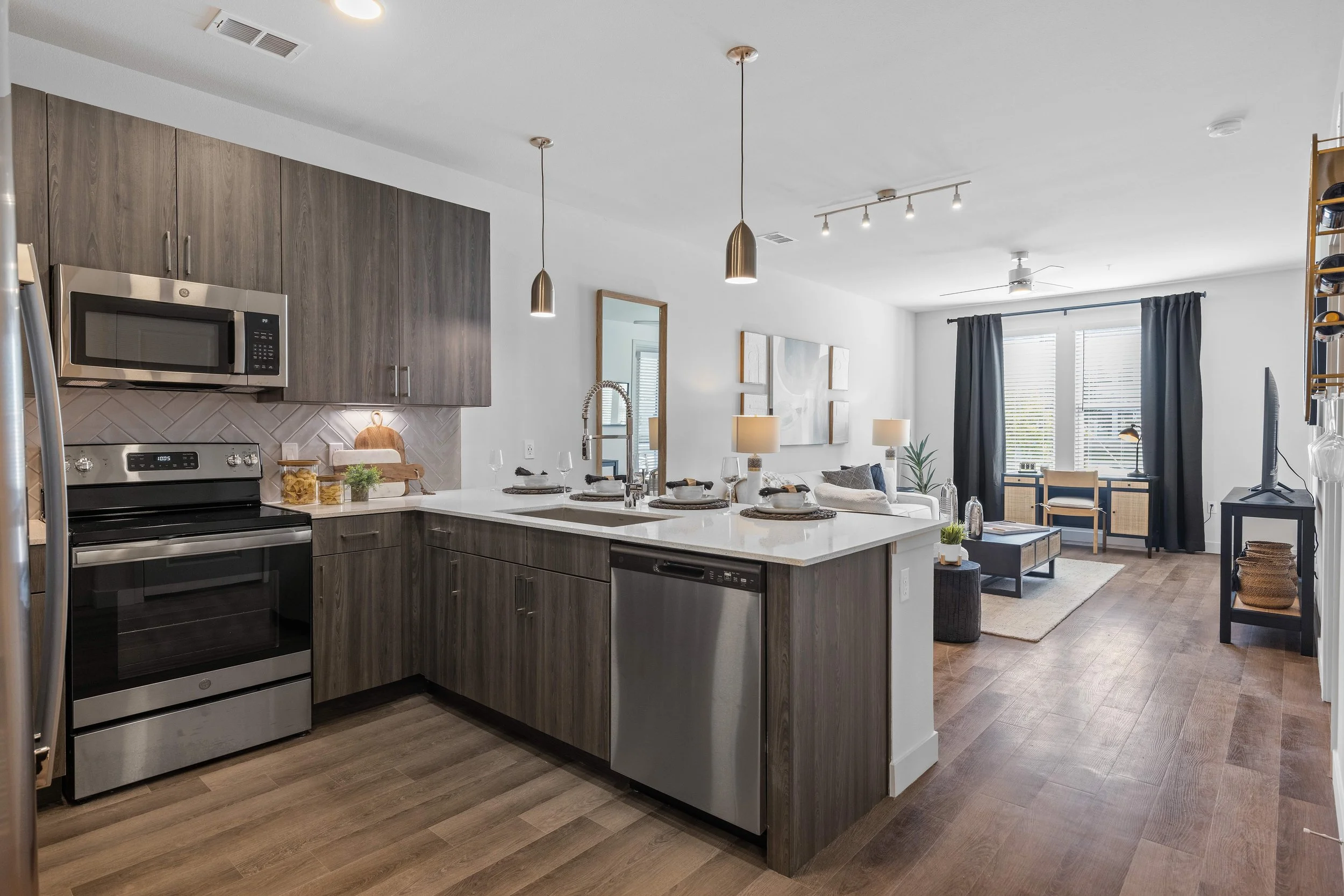 Open-concept kitchen and living room with wood cabinets, stainless steel appliances, island with place settings, and a seating area with lamps and artwork.