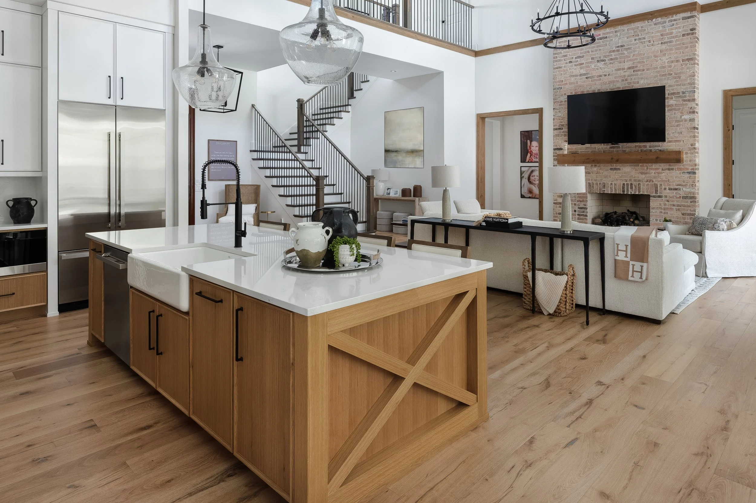Open-concept living room and kitchen with a high ceiling, brick fireplace with mounted TV, white furniture, wooden accents, and a staircase with a black metal railing.