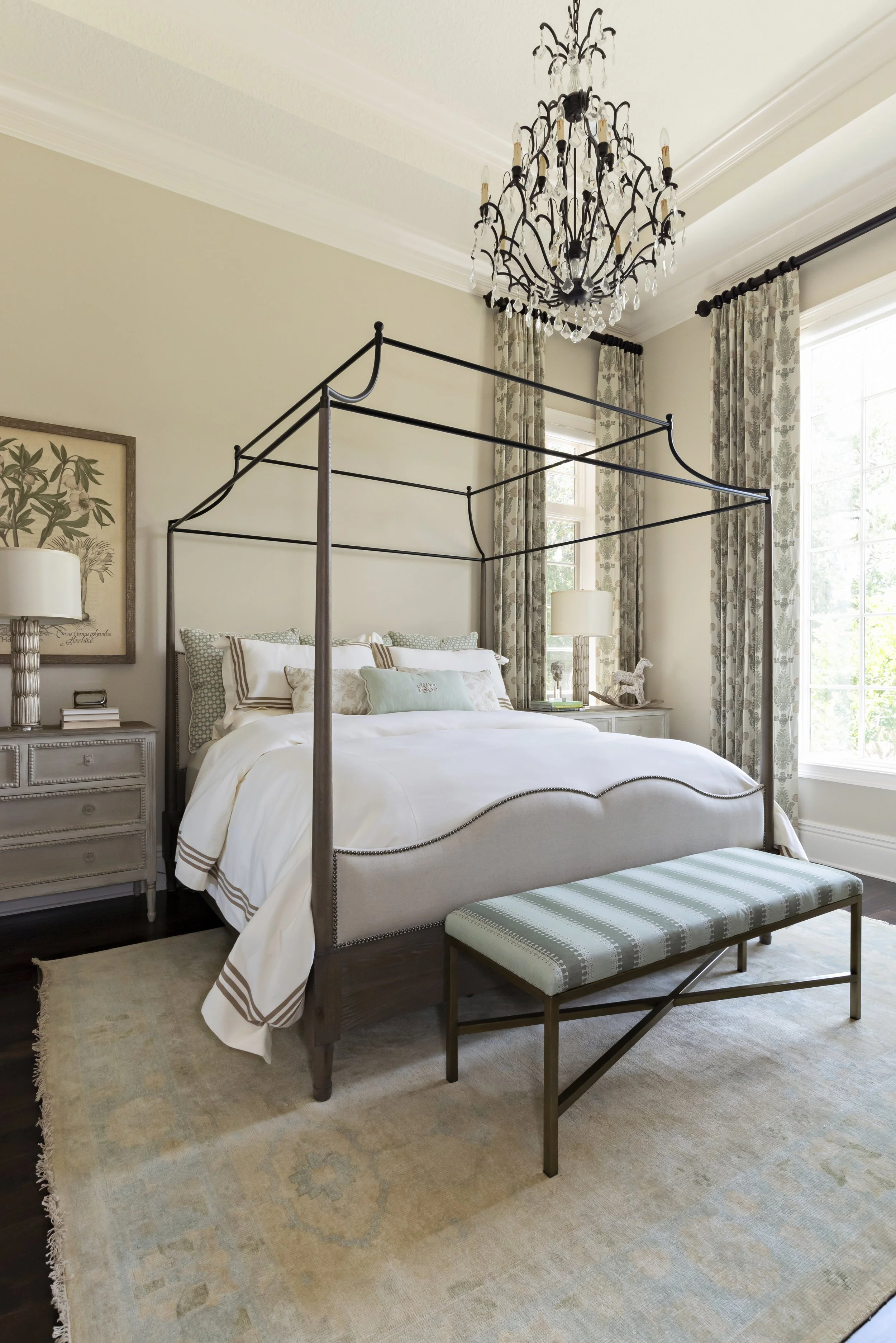 A bedroom with a bed that has a black metal canopy frame, white bedding with striped accents, and decorative pillows. There are two nightstands with lamps, a bench with a striped cushion at the foot of the bed, and large windows with floral curtains.