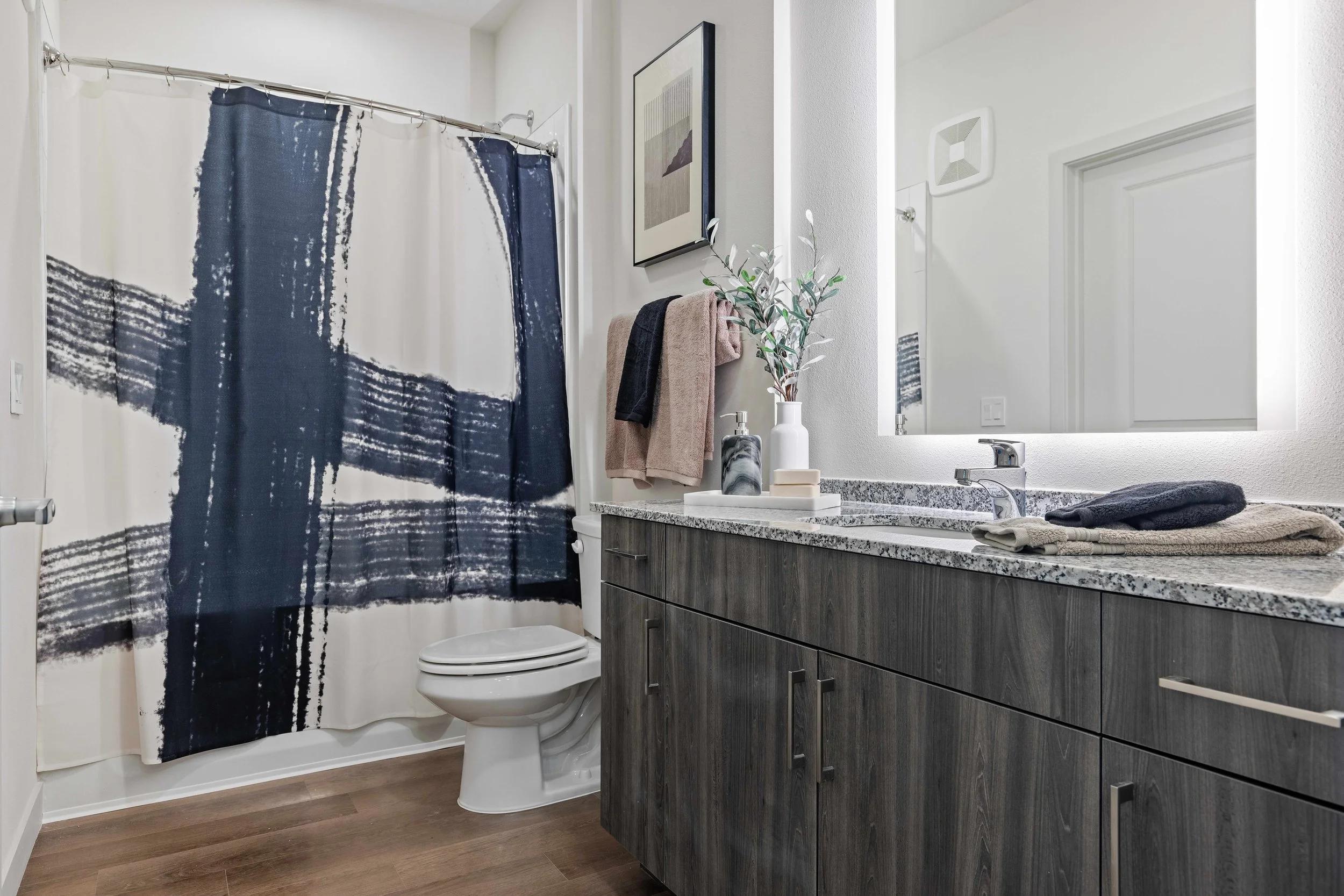 Bathroom with a white shower curtain with navy blue abstract patterns, a toilet, a vanity with a granite countertop, a mirror with backlighting, a potted plant, towels, and toiletries.