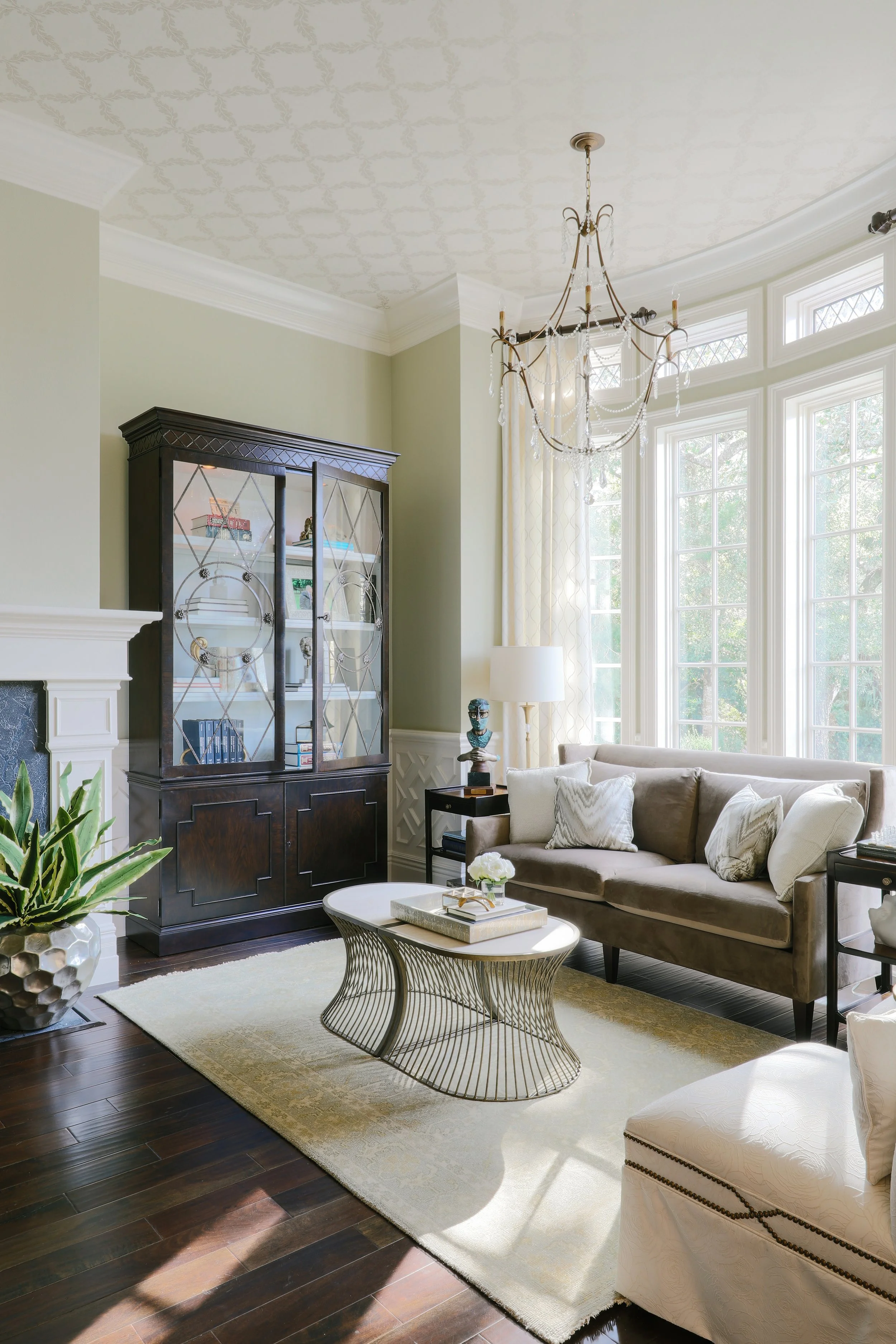 Bright living room with large windows, a chandelier, a beige sofa with pillows, a black display cabinet, a white and gold coffee table, a patterned rug, and a potted plant.