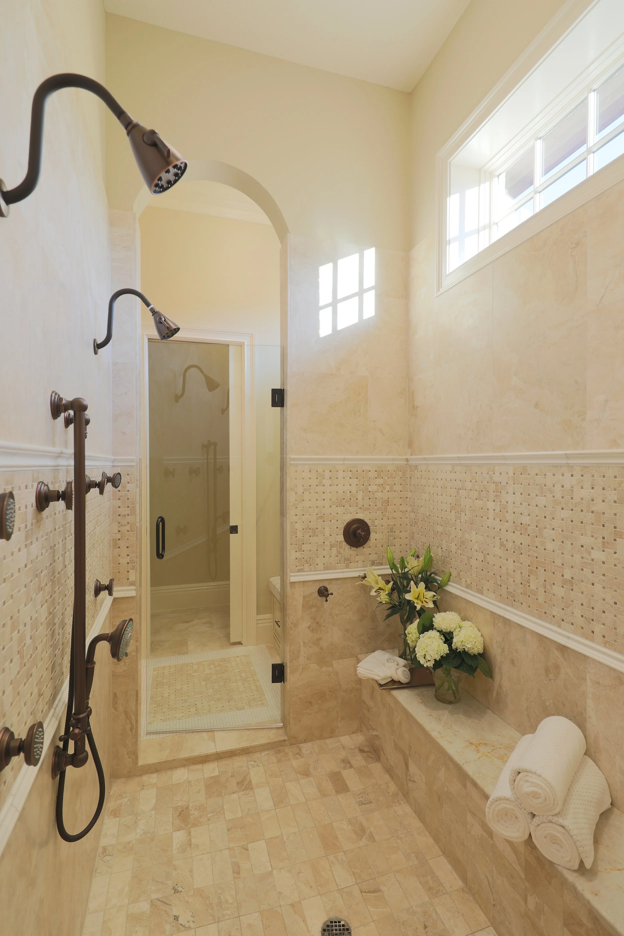 Bright and elegant bathroom with multiple shower heads, a glass shower door, a vase with white flowers, and neatly rolled white towels.