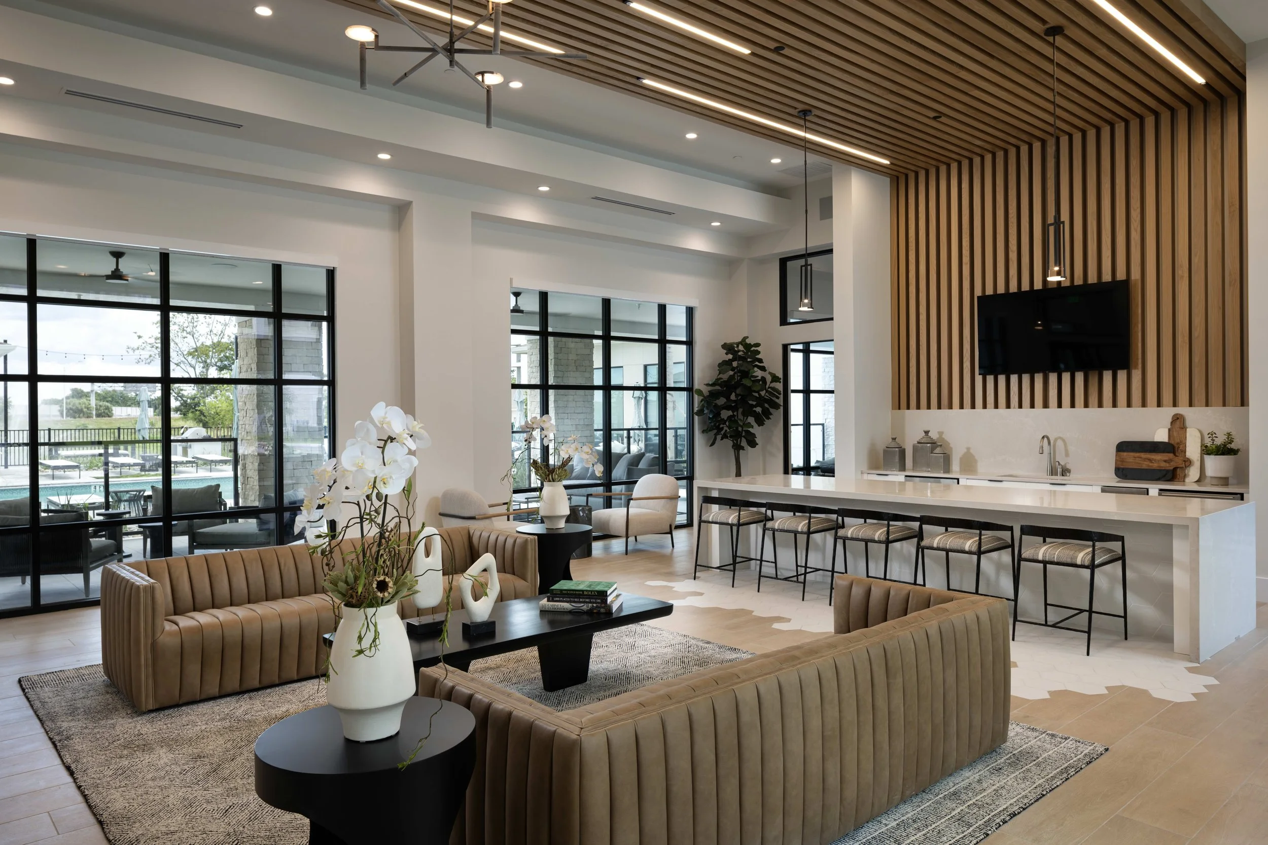 Modern open-concept living area with large windows, beige upholstered sofas, and a kitchen island with bar stools. Decor includes flowers in vases and hanging pendant lights.