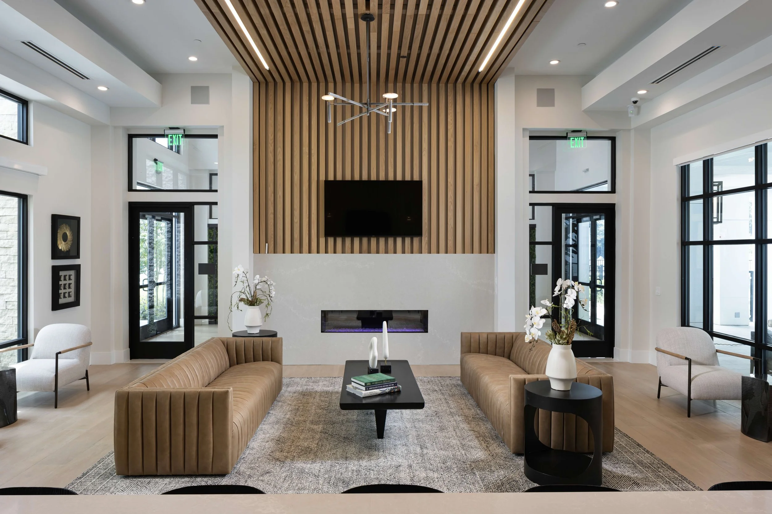Modern living room with a high ceiling, wooden panel accents, wall-mounted TV, fireplace, large windows, and contemporary furniture including two tan sofas and white armchairs, decorated with vases of white orchids and a black coffee table.