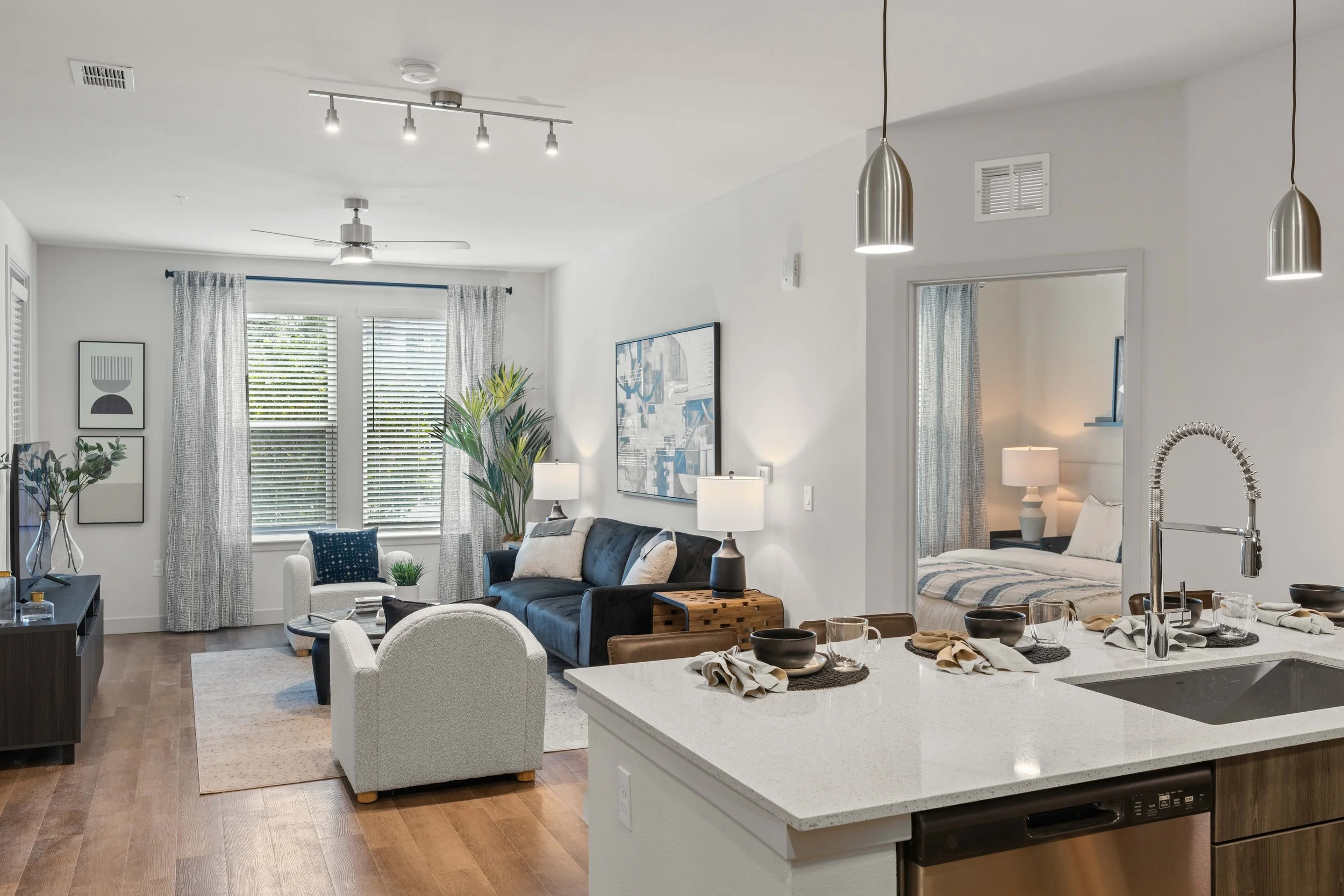 Bright, modern living space with large windows, curtains, and artwork. There is a white armchair, a gray sofa, and plants. The kitchen island with a sink and black bowls is visible in the foreground, and a dining table set with napkins and bowls is a