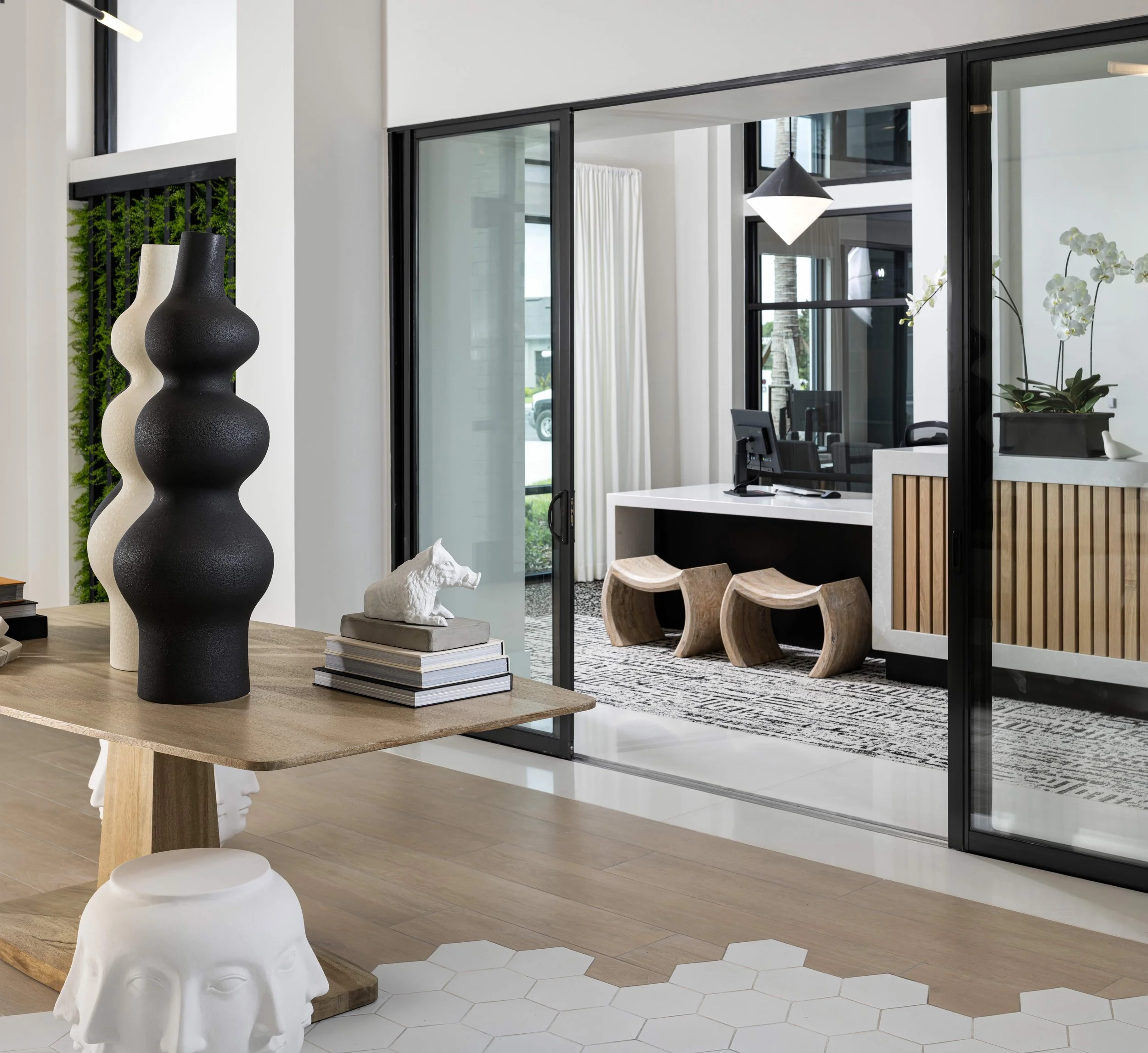 Modern interior with a wooden table, black and white vases, and sculptures. Glass sliding doors open to an outdoor area with a tropical tree in view.