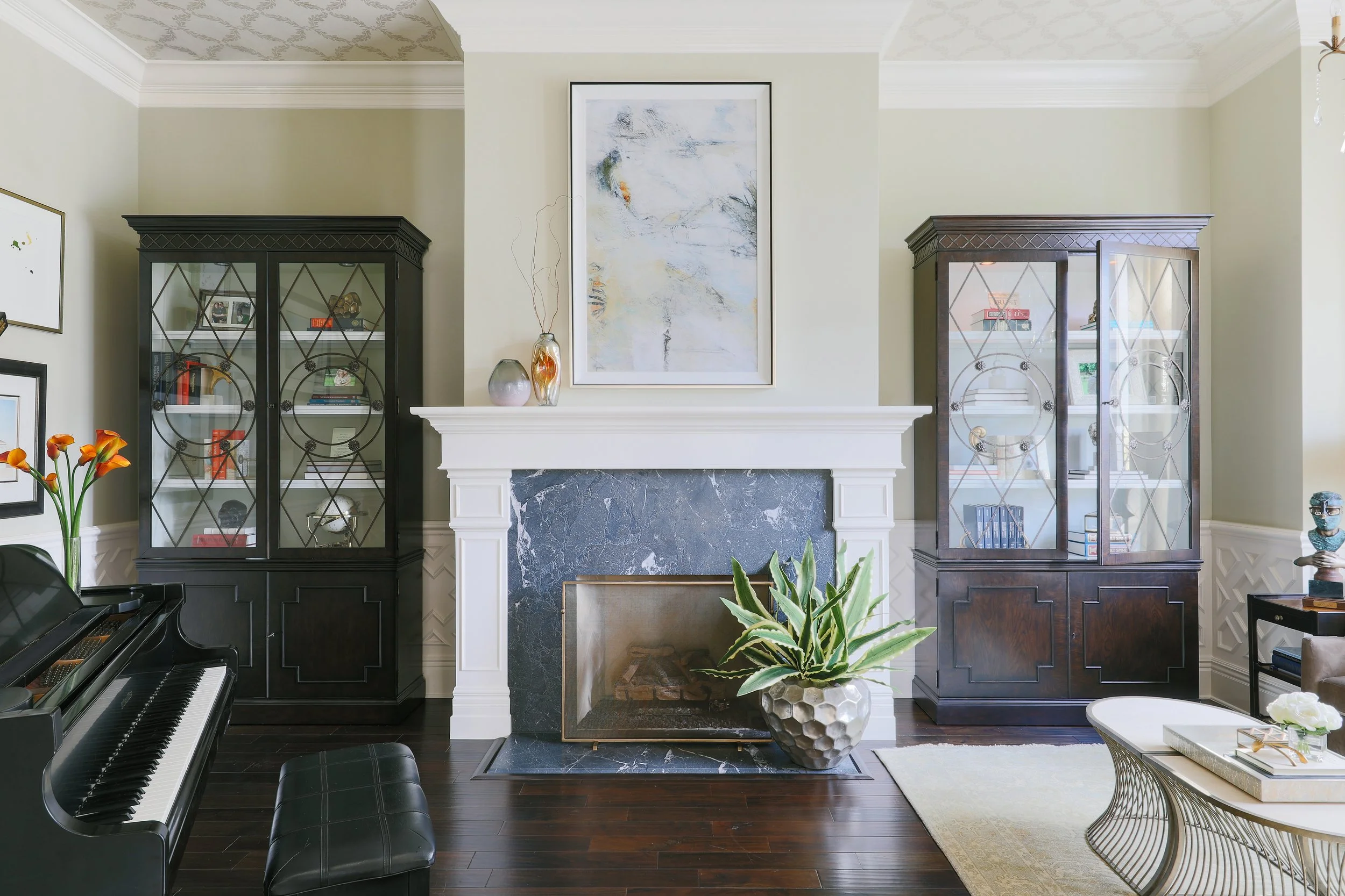 Living room with white fireplace, black and glass display cases, abstract wall art, potted plant, and a piano with bench.