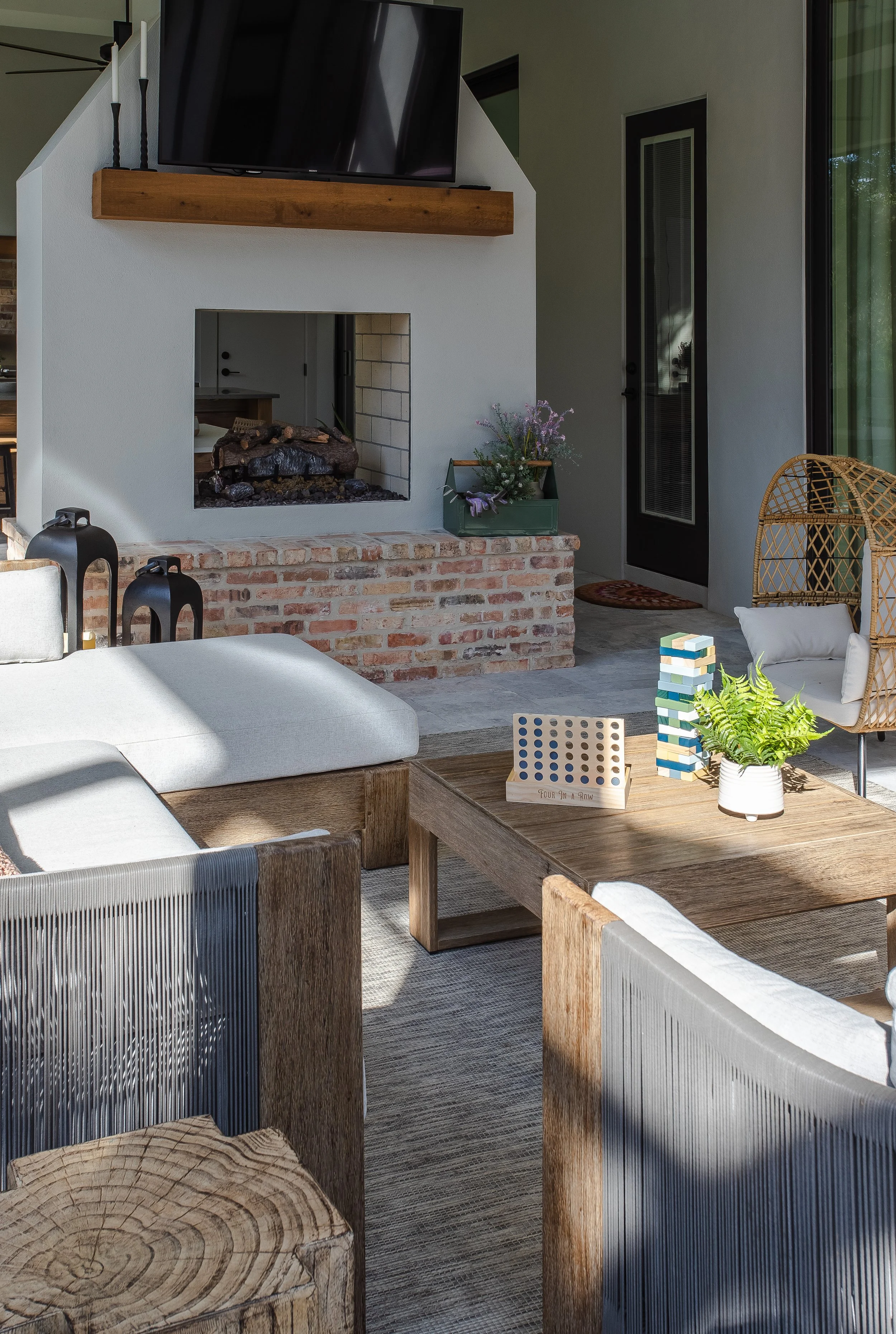 A cozy indoor living space with a fireplace, a wall-mounted TV above it, a wooden coffee table, and various seating options including a cushioned bench and wooden chairs. Decor includes a potted plant, a small decorative tower, and flowers placed nea