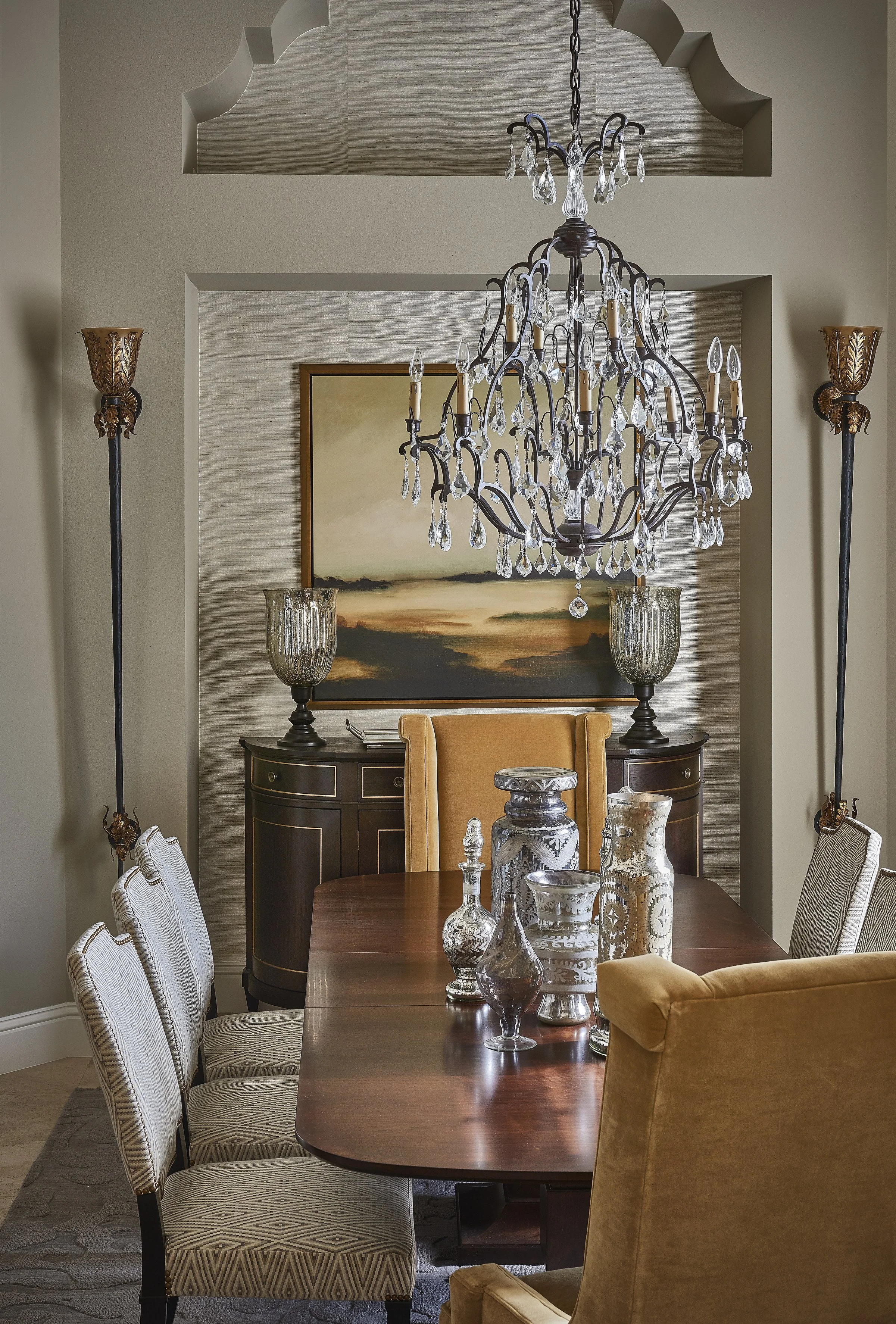 A dining room with a wooden table surrounded by upholstered chairs, a large chandelier hanging above, and decorative glass vases on the table. In the background, there's a sideboard with a landscape painting and two ornate glass lamps.