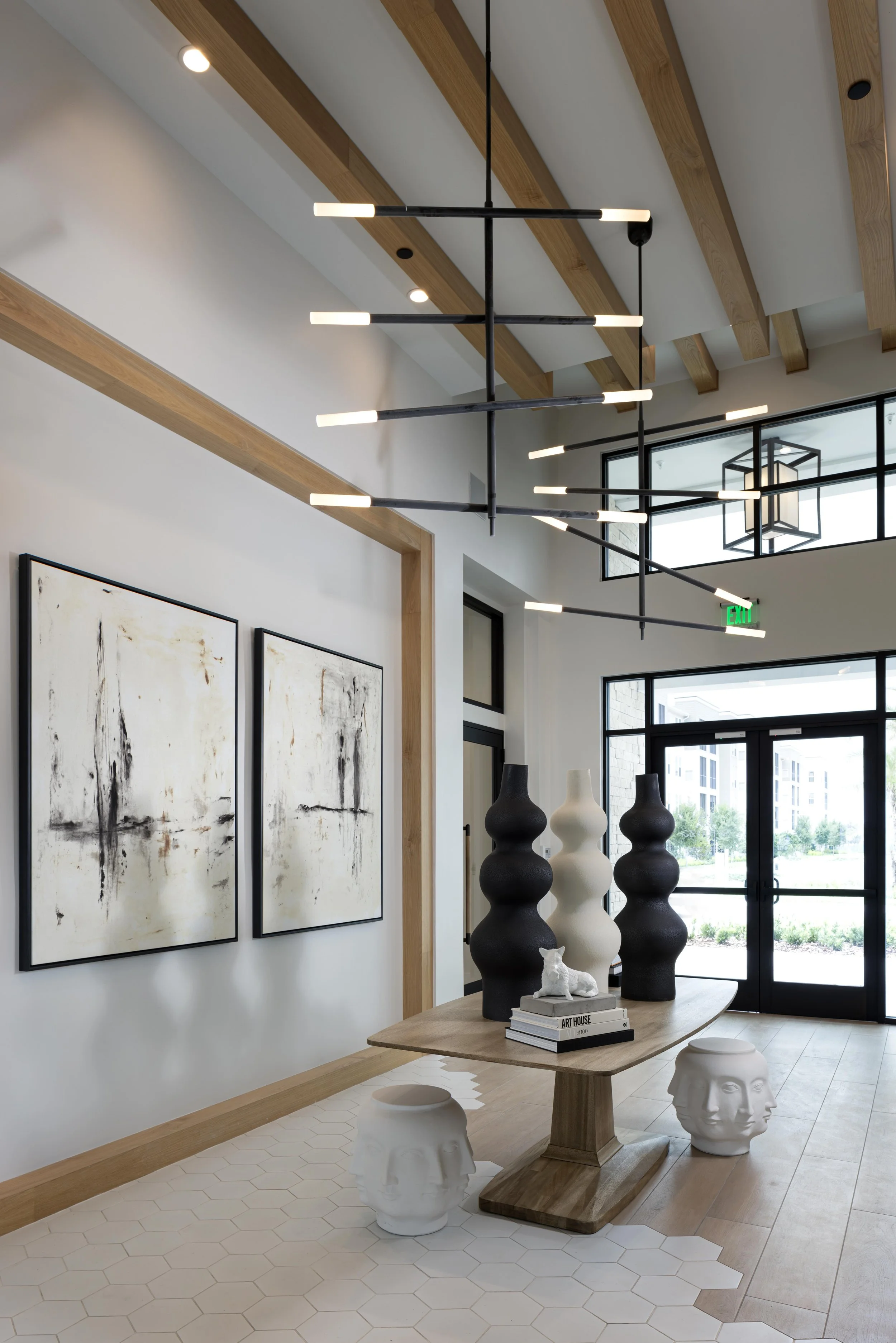 Modern interior space with a wooden dining table, black and white vases, art books, and decorative bust sculptures, with abstract artwork on the wall and large windows in the background.