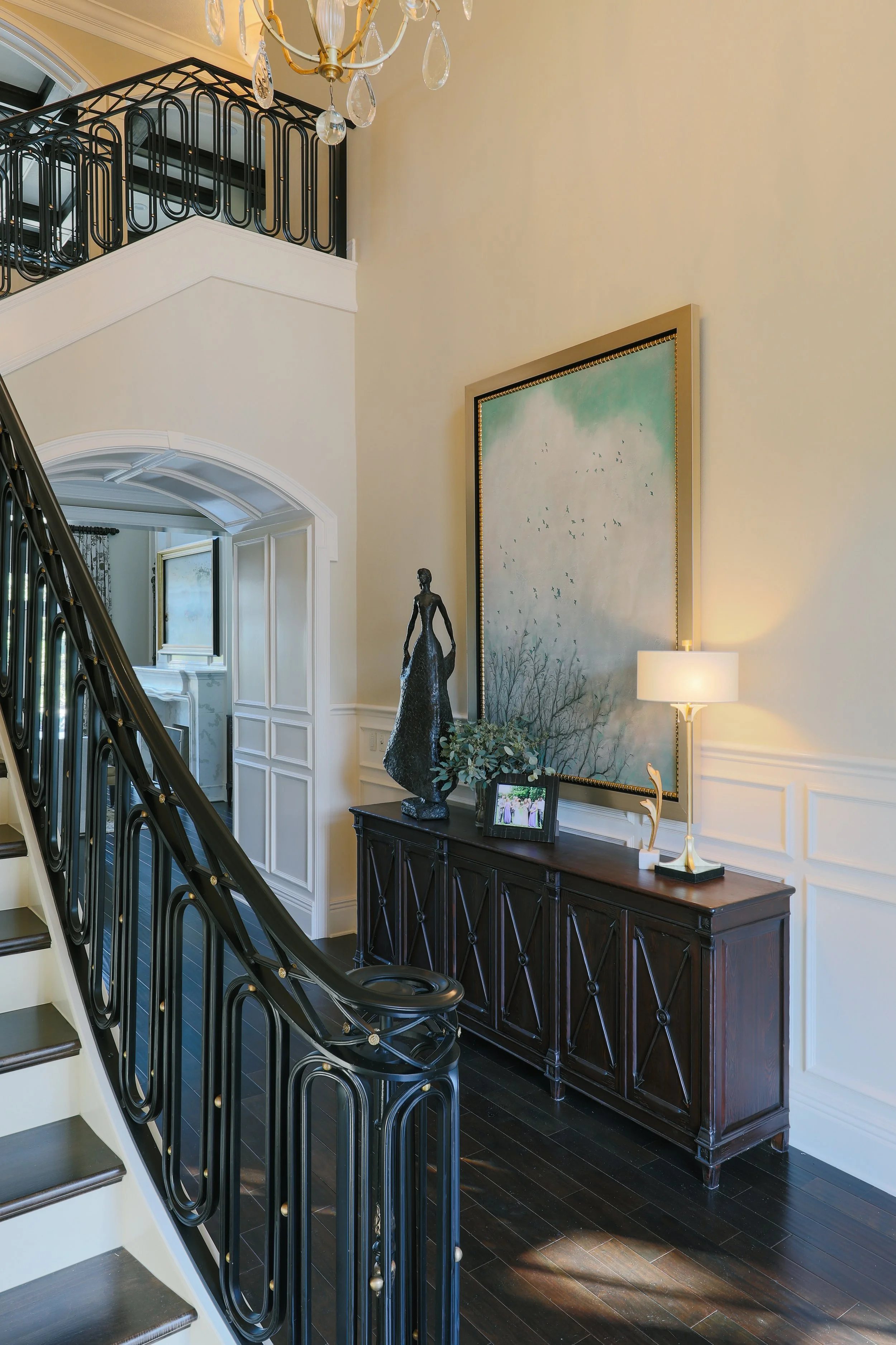 Interior of a home with a staircase, black iron railing, a dark wood console table, a large framed painting of a cloudy sky with birds, a small statue of a woman, a photo frame, a lamp, and a chandelier hanging from the ceiling.
