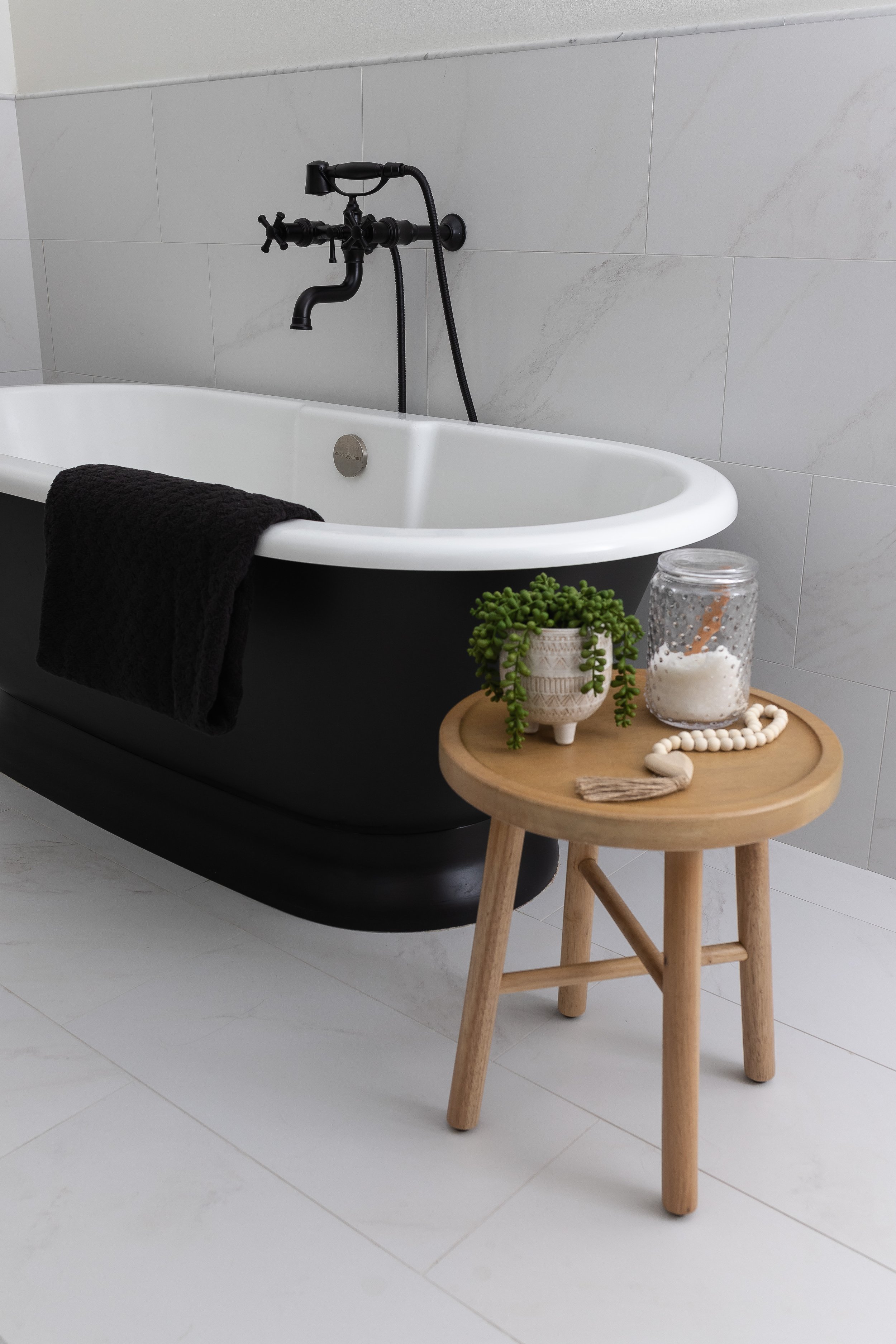 Bathroom with a black clawfoot tub, a wooden stool, a potted plant, a jar of bath salts, and a black towel.