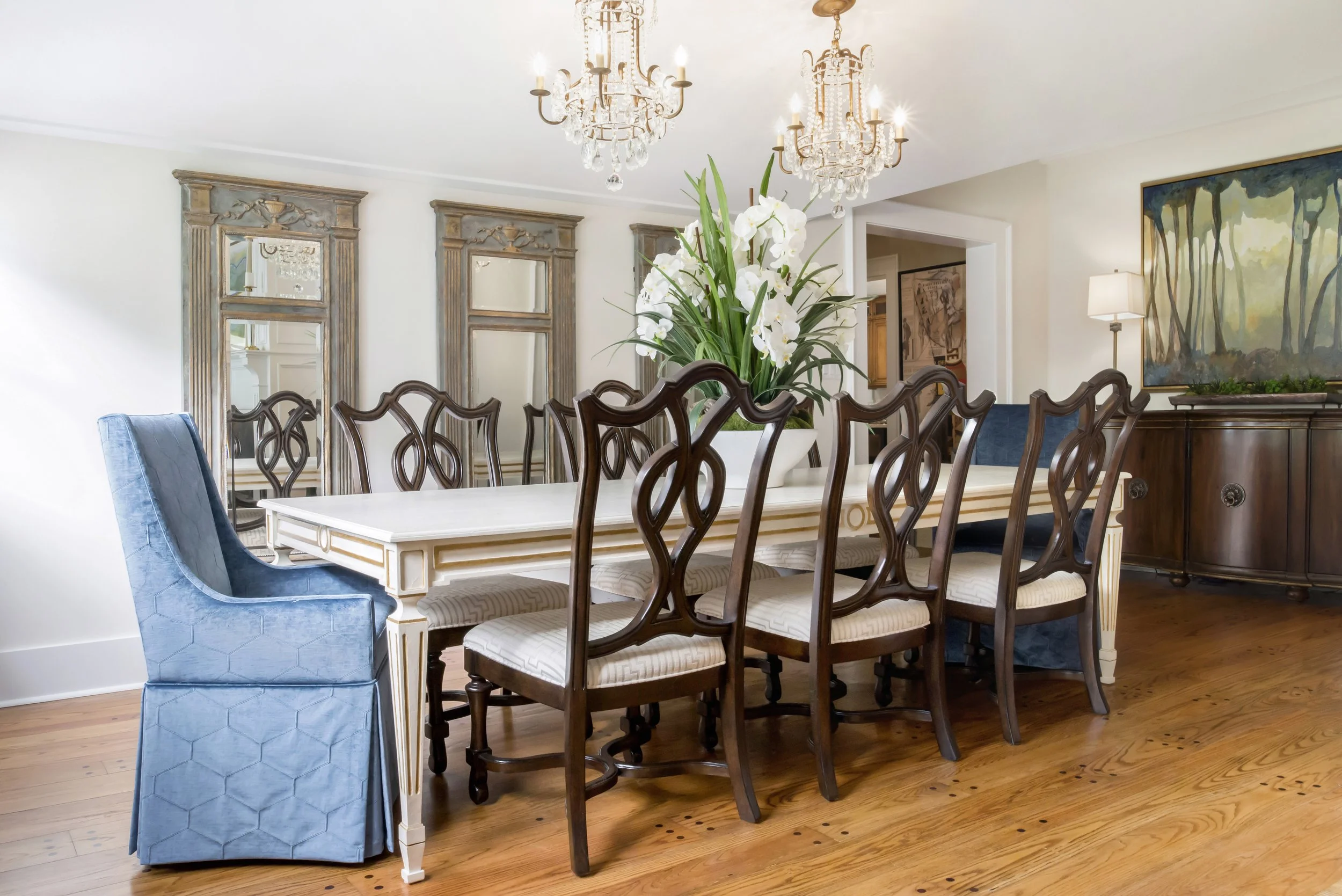 Dining room with a long white table, surrounded by wooden chairs, a blue upholstered armchair, and a large floral arrangement with white orchids in a white pot. There are three crystal chandeliers hanging from the ceiling, and three mirrors with orna