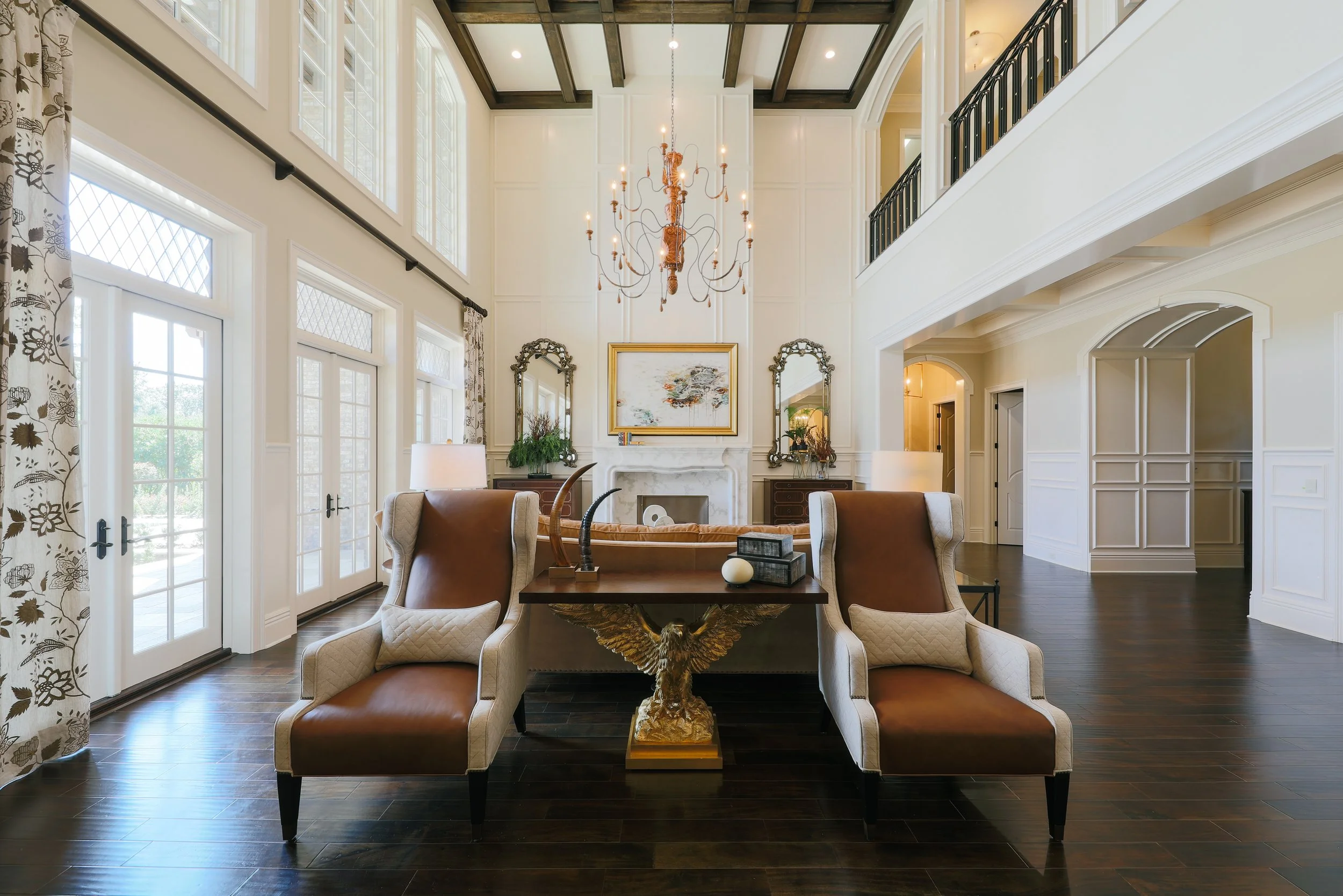 Interior of a spacious living room with high ceilings, large windows, a chandelier, fireplace, and elegant furniture.